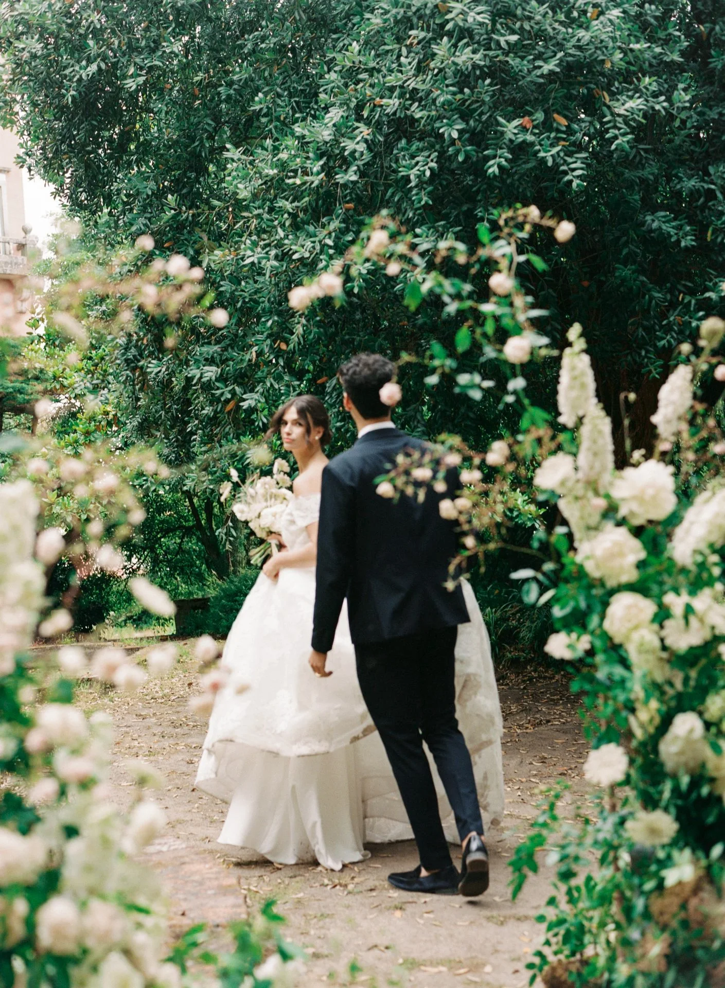 A bride and groom walking outdoors through a garden or park, surrounded by lush green foliage and flowering plants, during their wedding.