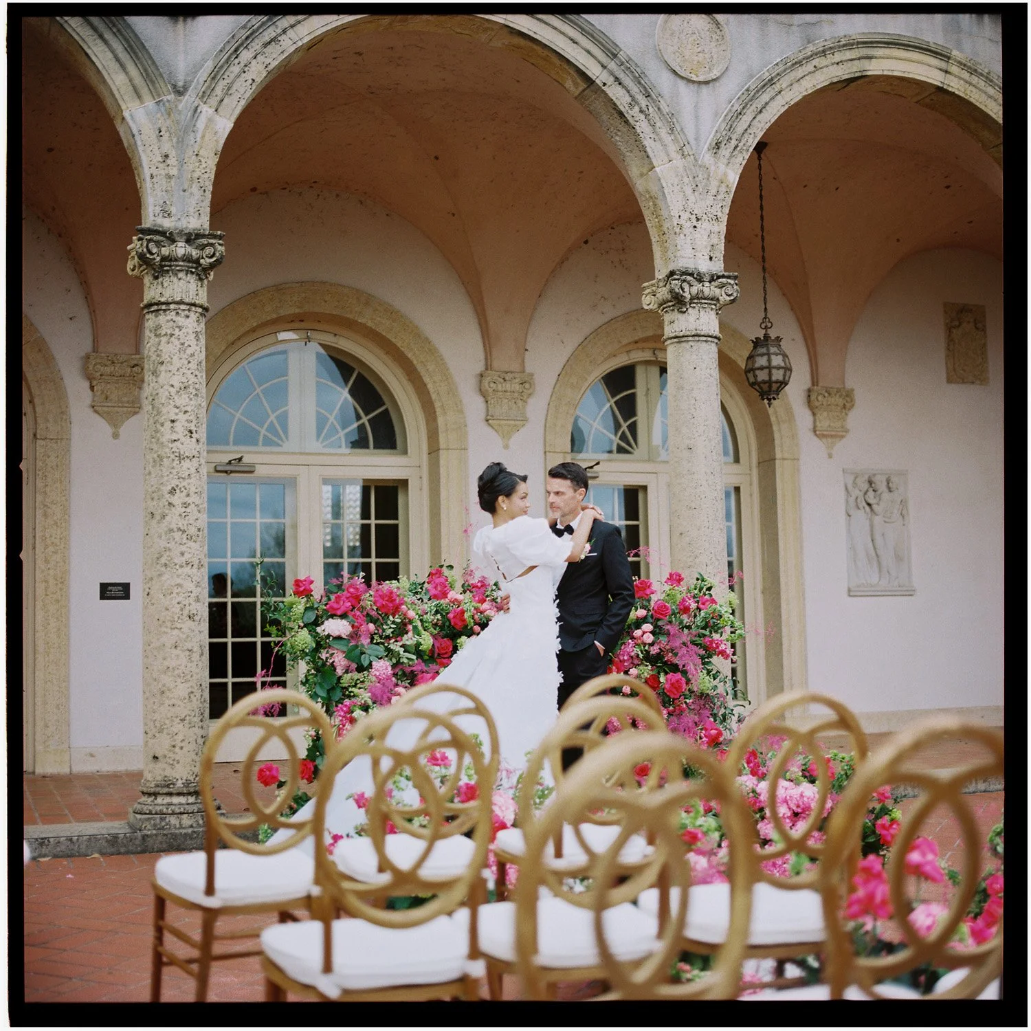 A bride and groom dancing at a wedding, with flower arrangements and classical architecture in the background.