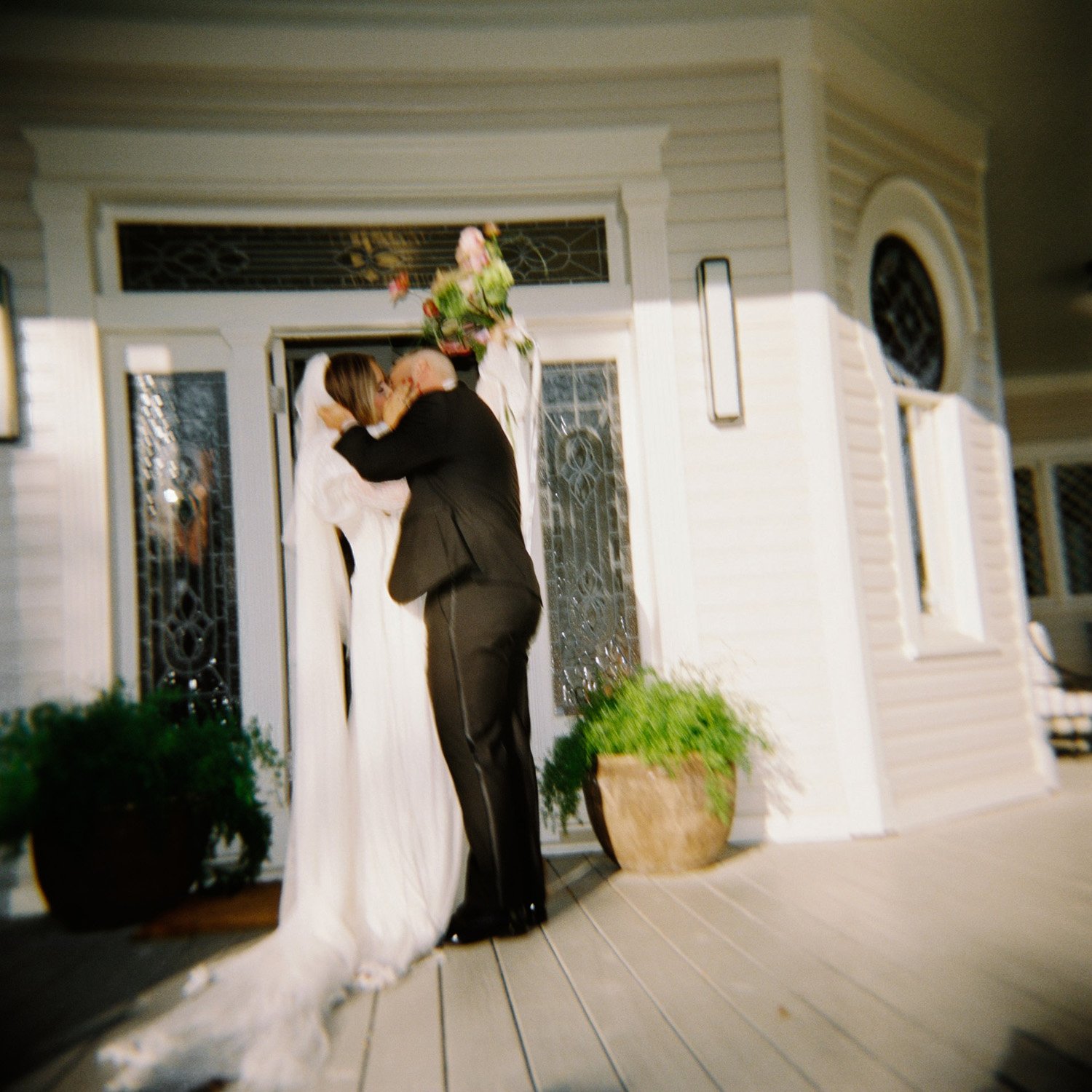 A couple dressed in wedding attire kissing outside a house. The woman is wearing a wedding gown and veil, and the man is in a tuxedo. The woman is holding a bouquet of flowers above their heads.