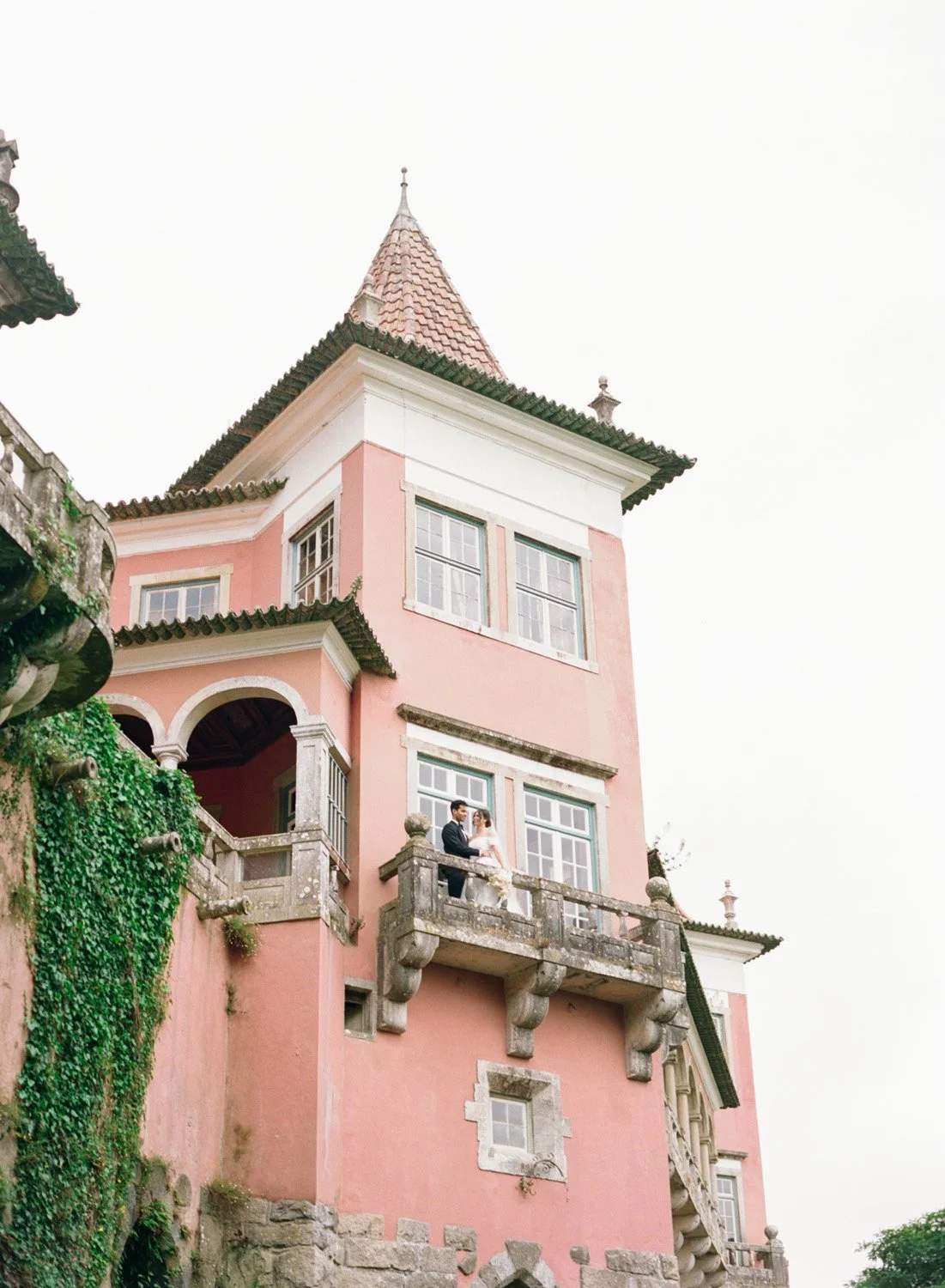 A pink multi-story castle-like house with a turret, green tiled roof, and large windows. Two people, a man and a woman dressed in formal attire, are standing on a small balcony on the second floor.