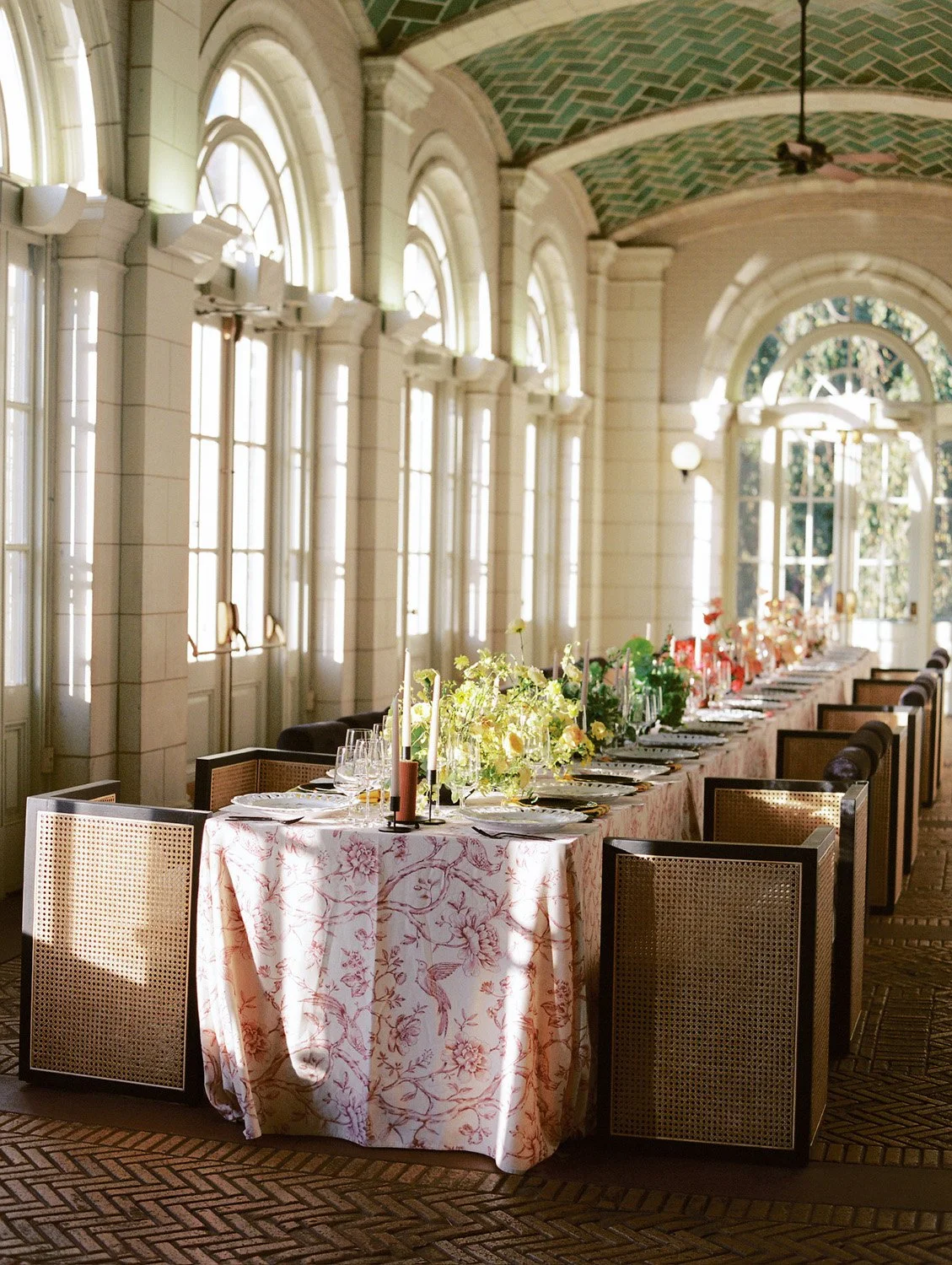 Elegant banquet table set with floral centerpieces, candles, and fine dinnerware along a sunlit arched windowed wall in a grand room.