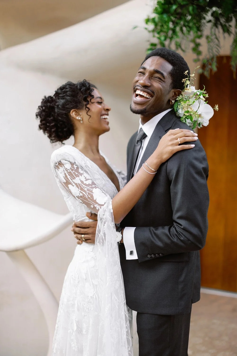 A happy couple on their wedding day, dancing and laughing indoors. The bride wears a white lace wedding dress with floral embroidery, and the groom wears a black suit with a white shirt and black tie. The bride's hand rests on the groom's shoulder, and they are both smiling joyfully.