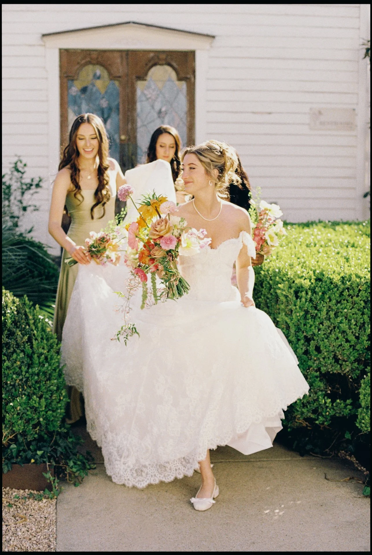 A bride in a white wedding dress holding a bouquet of pink and yellow flowers, walking outdoors with bridesmaids in matching dresses behind her.