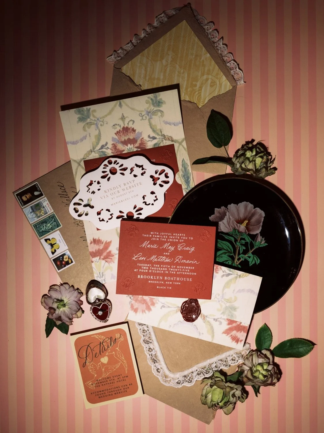 Wedding stationery and decor with floral accents, including invitations, RSVP card, a black plate with a floral design, a black flower, and pink and green flowers, set on pink striped background.