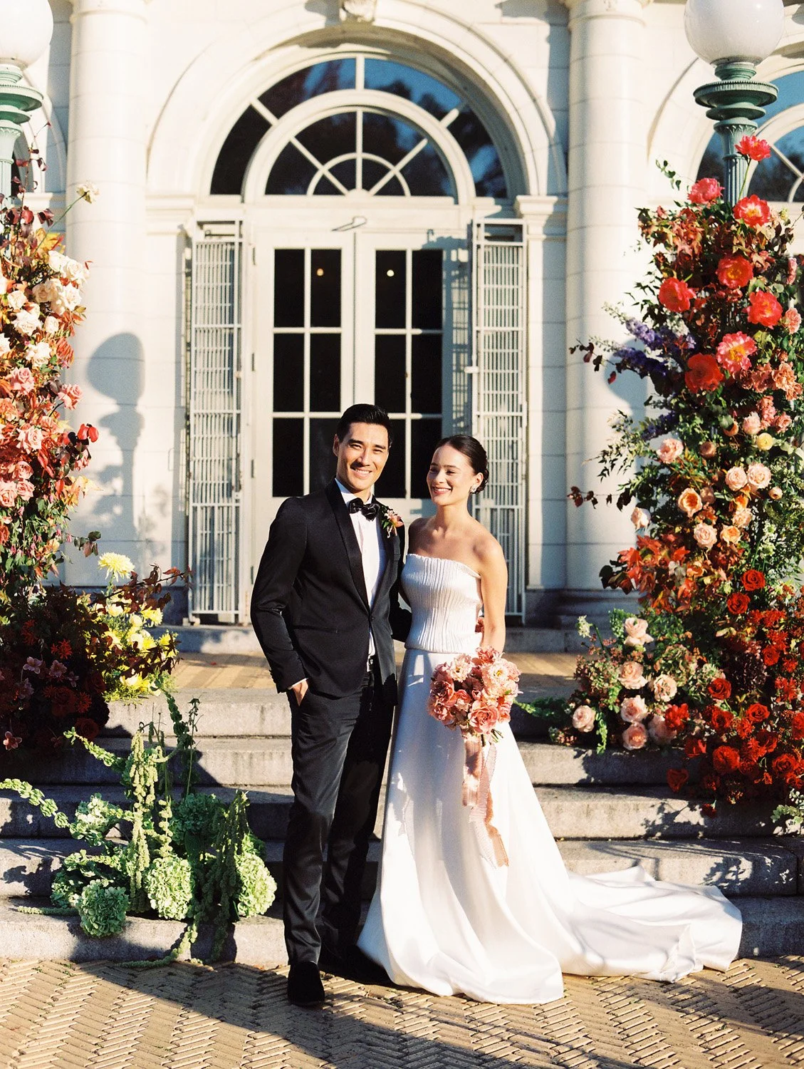 A newlywed couple standing on steps outside a white building with large windows, surrounded by colorful flowers. The groom is in a black tuxedo, and the bride is in a strapless white wedding gown holding a bouquet of pink and peach flowers.