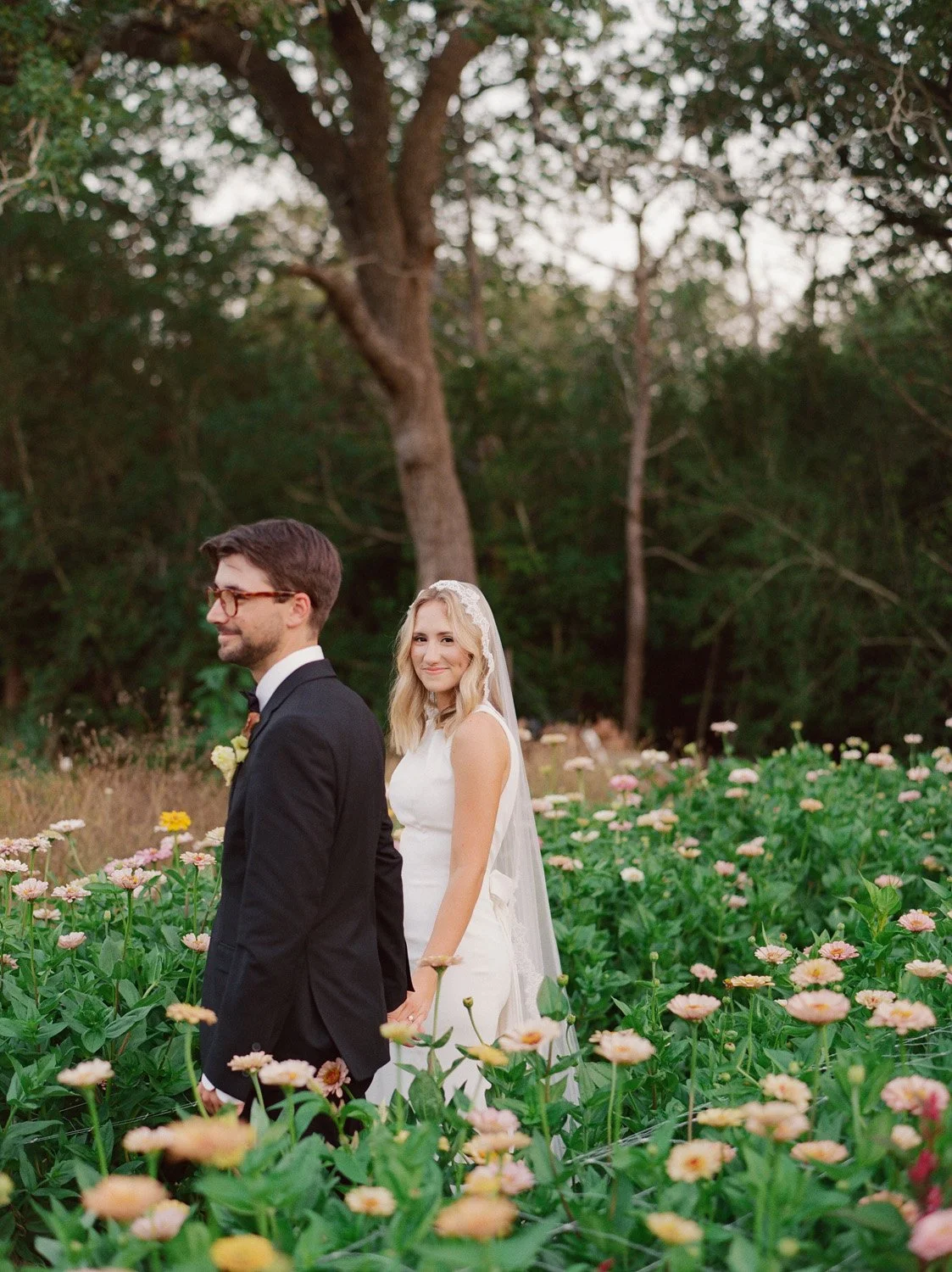 Bride and groom standing outdoors in a field of pink and yellow flowers, with trees in the background, during their wedding photoshoot, holding hands behind their backs.