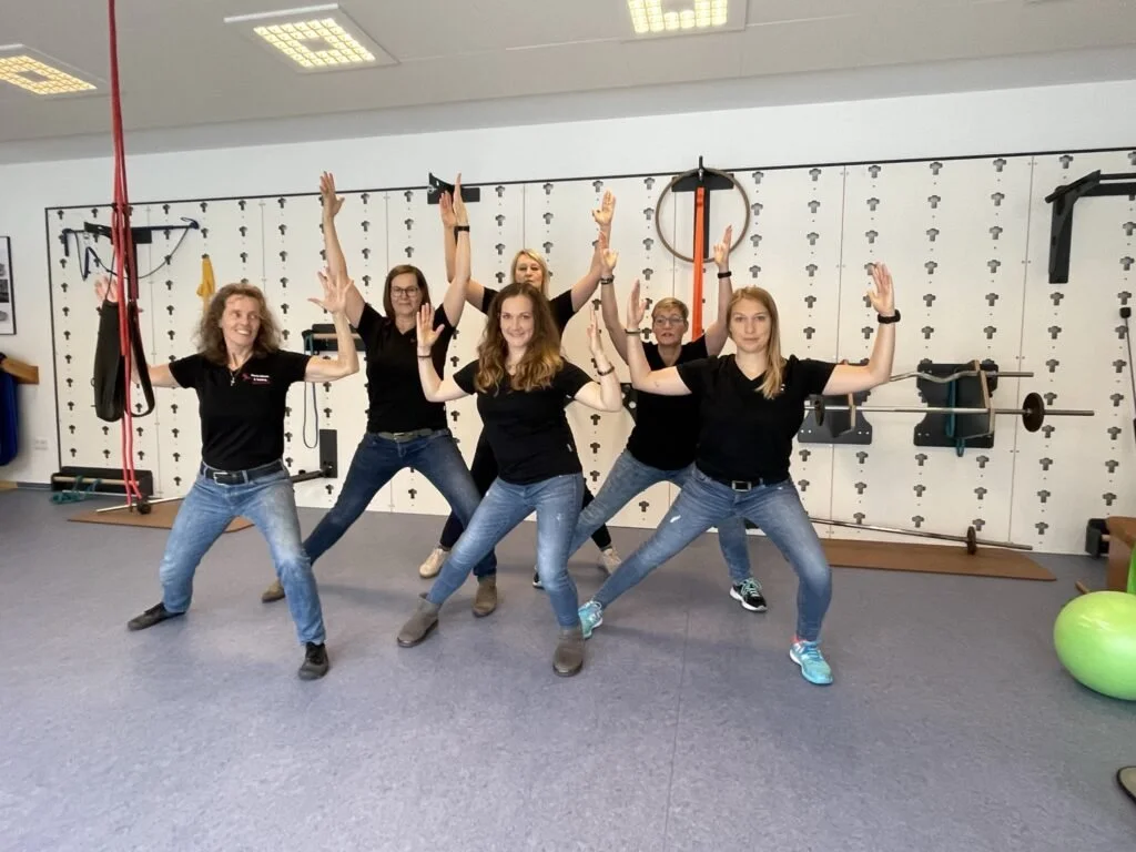 Fünf Frauen in sportlicher Kleidung posieren in einem Fitnessraum mit ausgestreckten Armen und lachend.