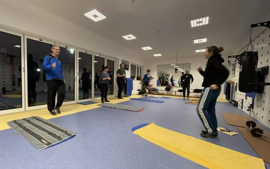 Menschen beim Fitness im Fitnessstudio, einige auf Yoga- oder Trainingsmatten, andere stehen und sprechen, mit Spiegeln an der Wand und Trainingsequipment im Hintergrund.