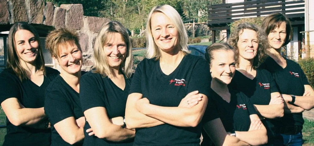 Gruppe von acht Frauen in schwarzen T-Shirts, die vor einem Außenbereich mit Steinen und Holzstrukturen stehen, lächelnd in die Kamera schauend.