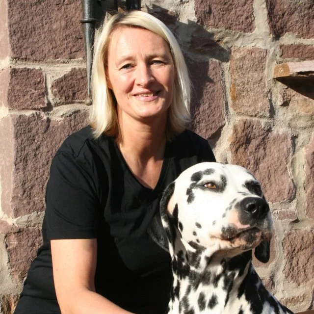 Eine blonde Frau in Schwarz sitzt neben einem Dalmatiner-Hund vor einer Steinmauer.