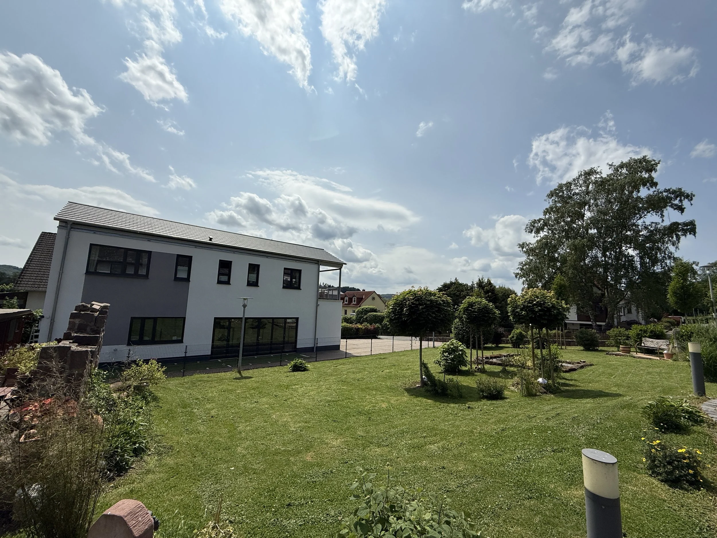 Ein Garten mit grünem Rasen, Bäumen, Blumen und einem modernen weißen Haus im Hintergrund bei sonnigem Himmel mit manchen Wolken.