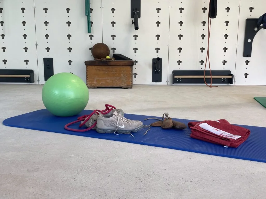 Trainingsausrüstung auf einer blauen Yogamatte, inklusive einem großen grünen Ball, weißer Turnschuhe, Seilen, Lederhandschuhen und einem roten Handtuch, in einem Fitnessraum.