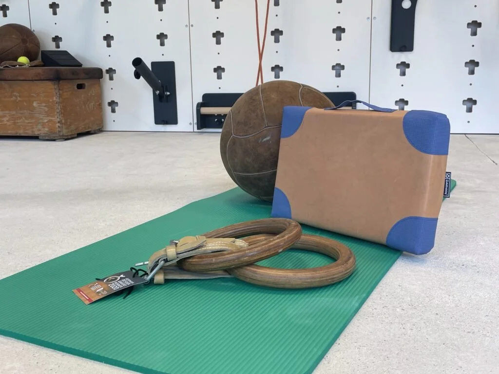 Fitnessgeräte wie Gymnastikmatte, Holzringen, Medizinball, Kasten und Wandmontage für Klimmzüge in einem Trainingsraum.