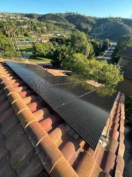 Solar panels installed on a tiled rooftop with a scenic view of rolling hills, trees, and a clear blue sky in the background.