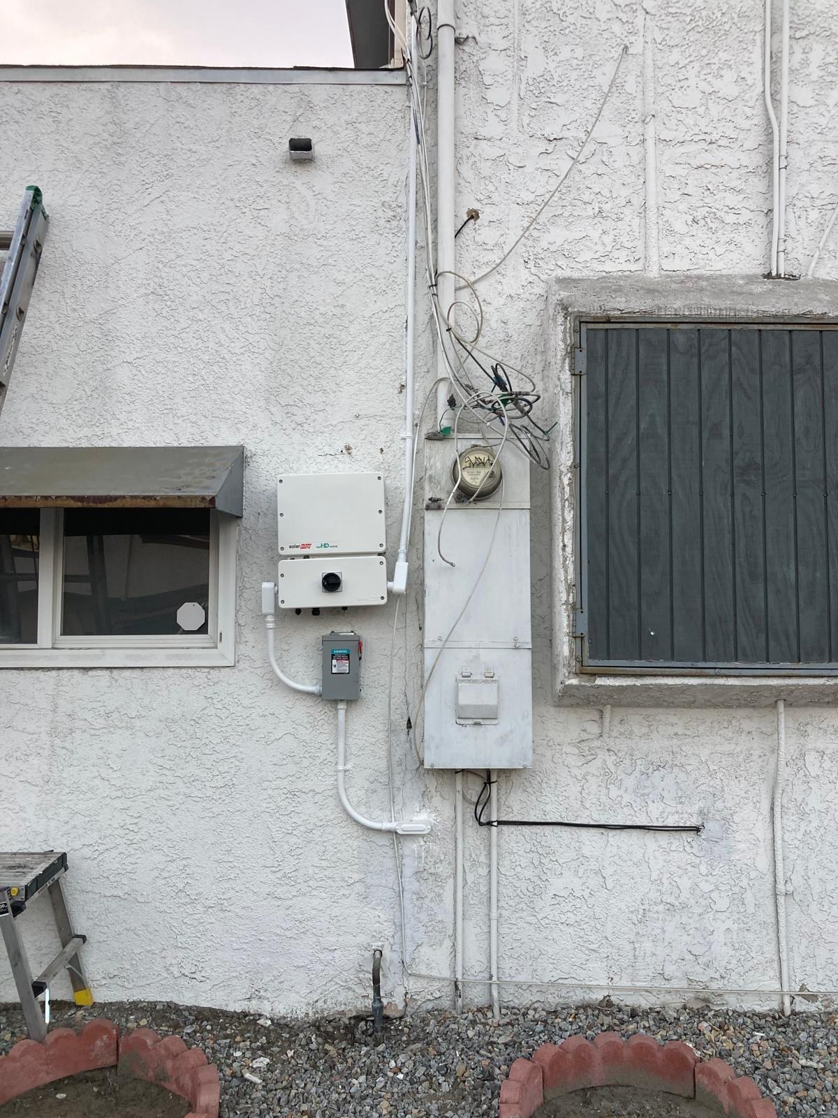 The exterior of a white stucco building features various utility installations, including an electric meter, utility boxes, and exposed cables, with two windows, one with black metal bars, and a ladder leaning against the wall.