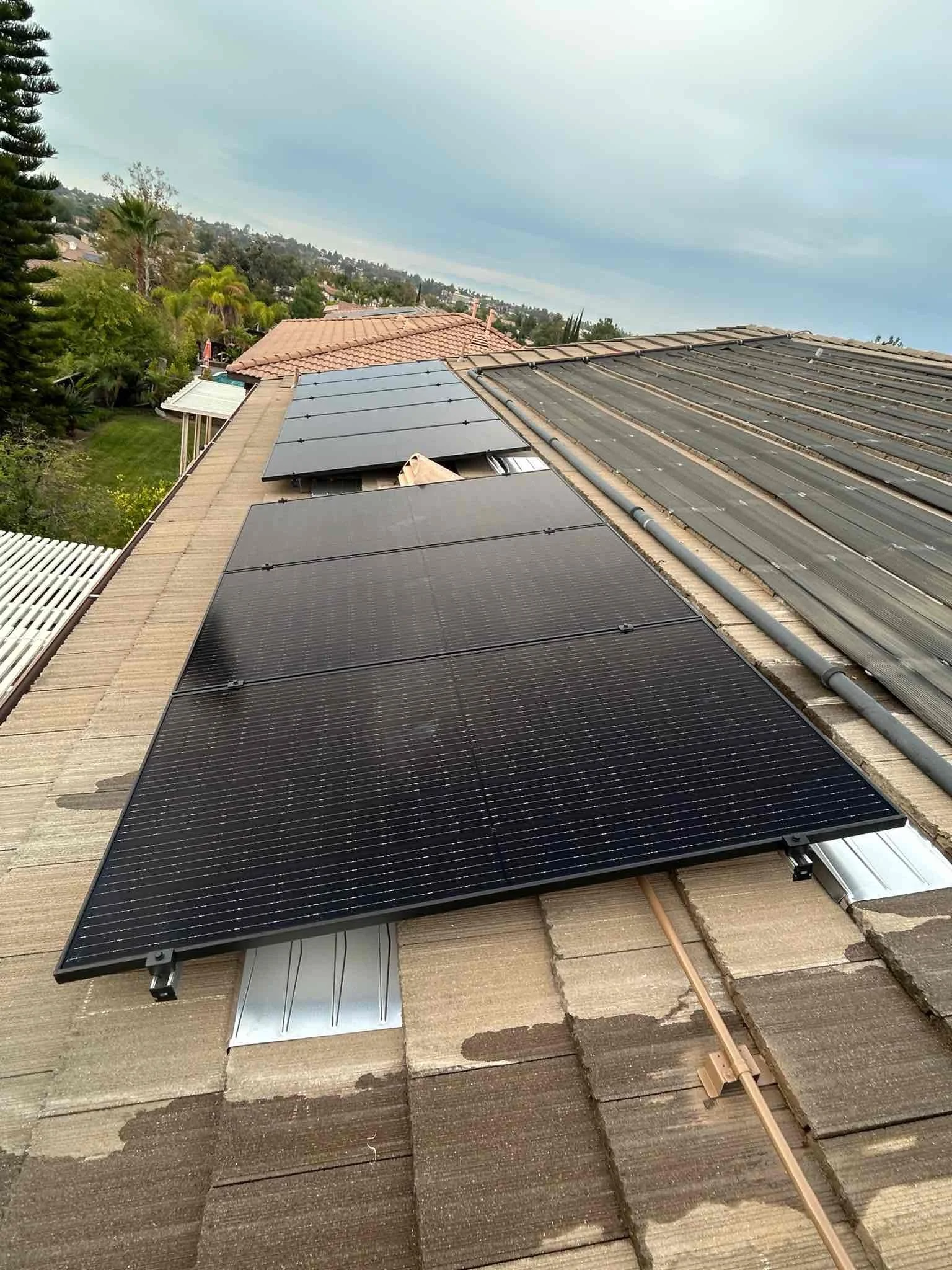 A rooftop with multiple solar panels installed on it, with some tiles visible. There are trees and other houses in the background under an overcast sky.