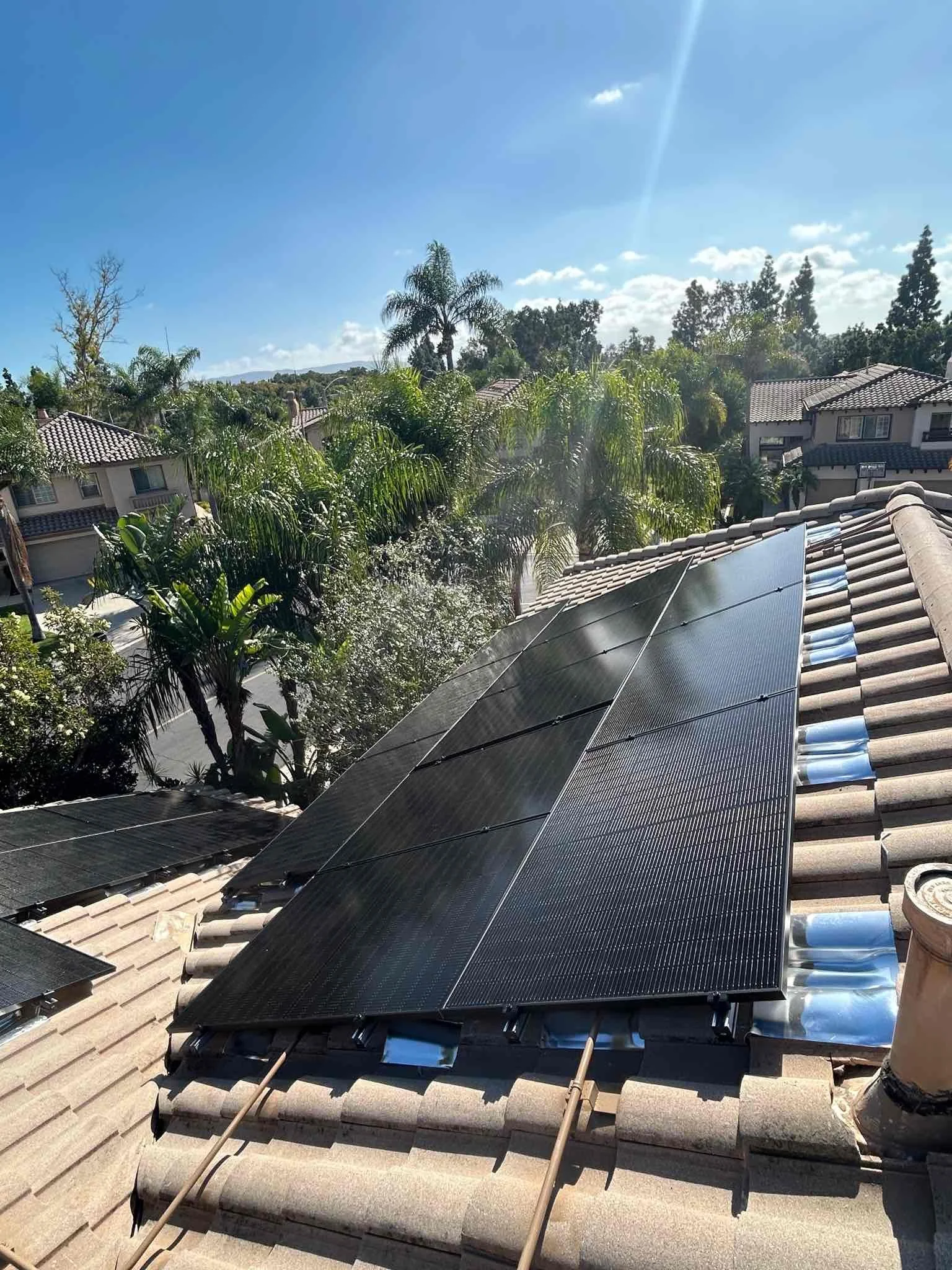 Solar panels installed on a tiled roof of a house, with trees and houses visible in the background under a clear blue sky.