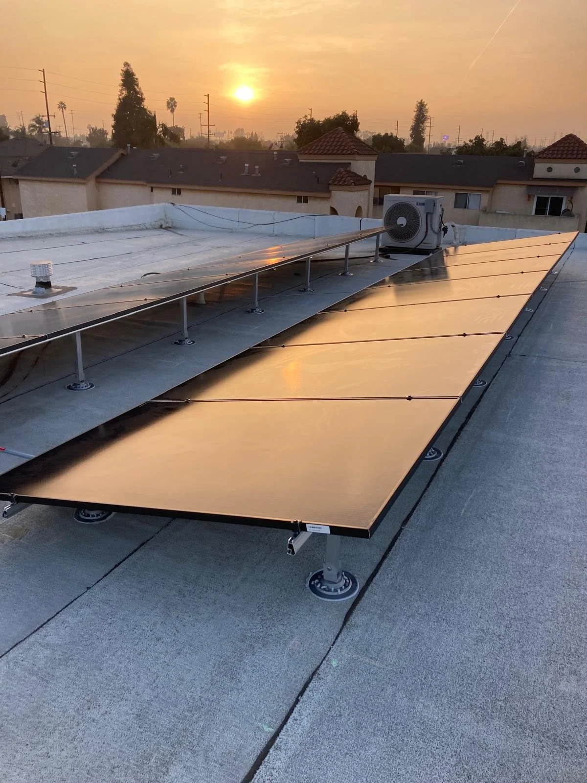 Sunset over a rooftop with solar panels and an air conditioning unit, with houses and trees in the background.