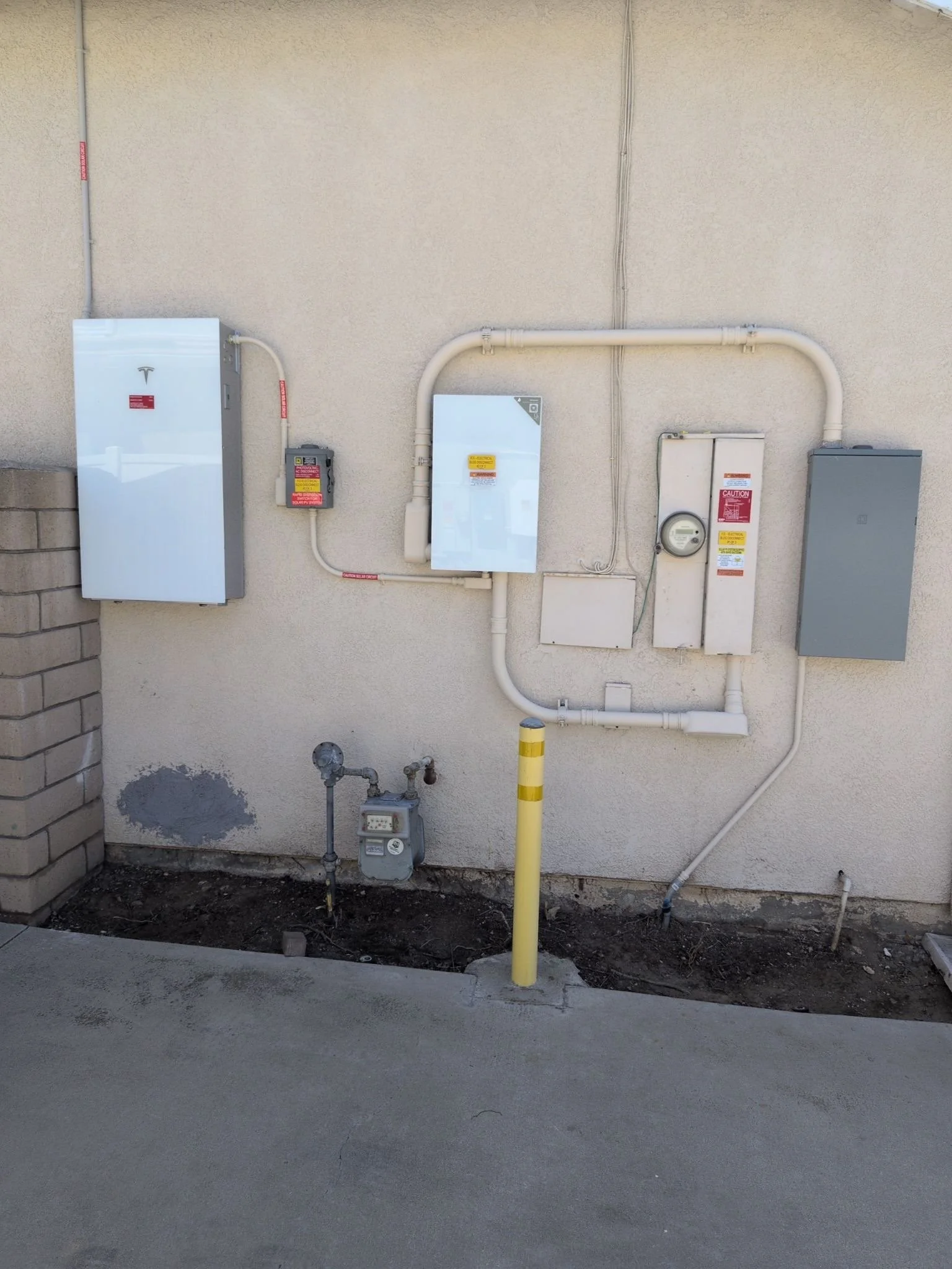 Outdoor wall with electrical boxes, pipes, and conduit. Yellow safety pole and some exposed soil and concrete ground in front.