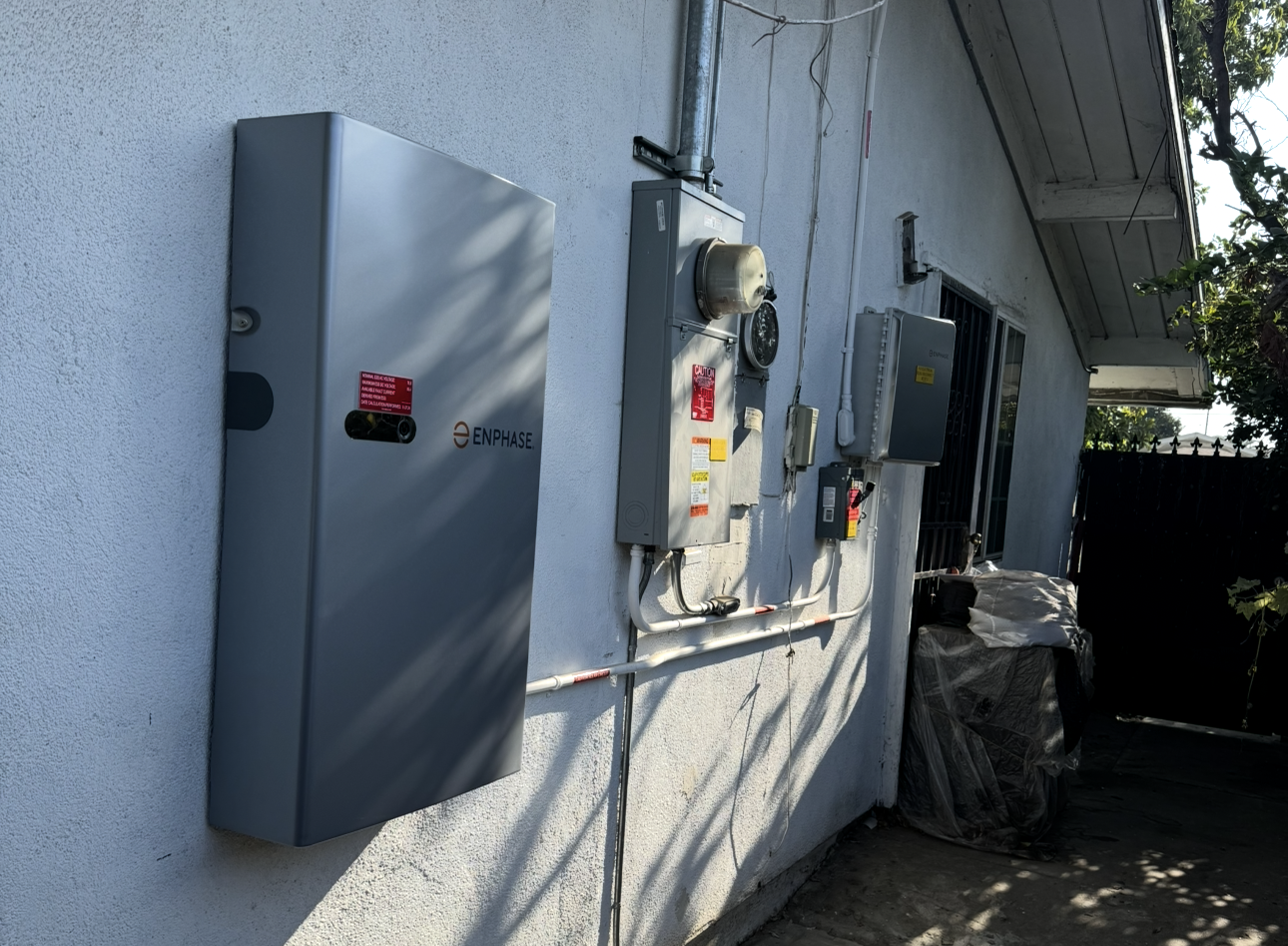 Exterior view of an electrical utility wall with various electrical boxes, meters, and panels mounted on a white wall under a roof overhang.