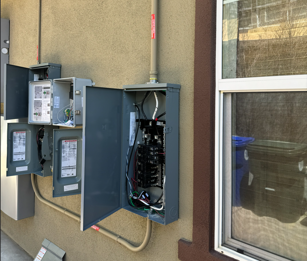 Open electrical meter and circuit breaker boxes mounted on a beige wall outside a building, with some wires and cables visible inside.