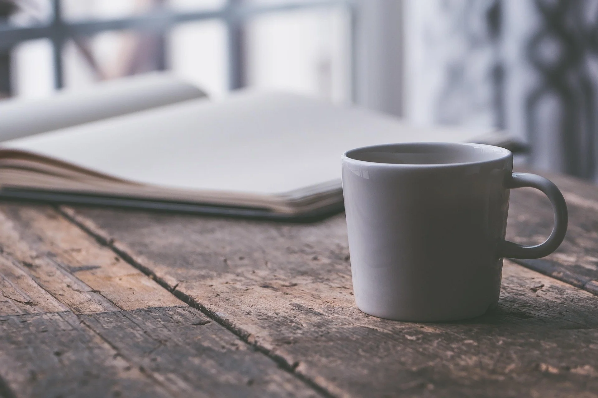 A white coffee mug and an open book sit on a rustic wooden table in natural light.
