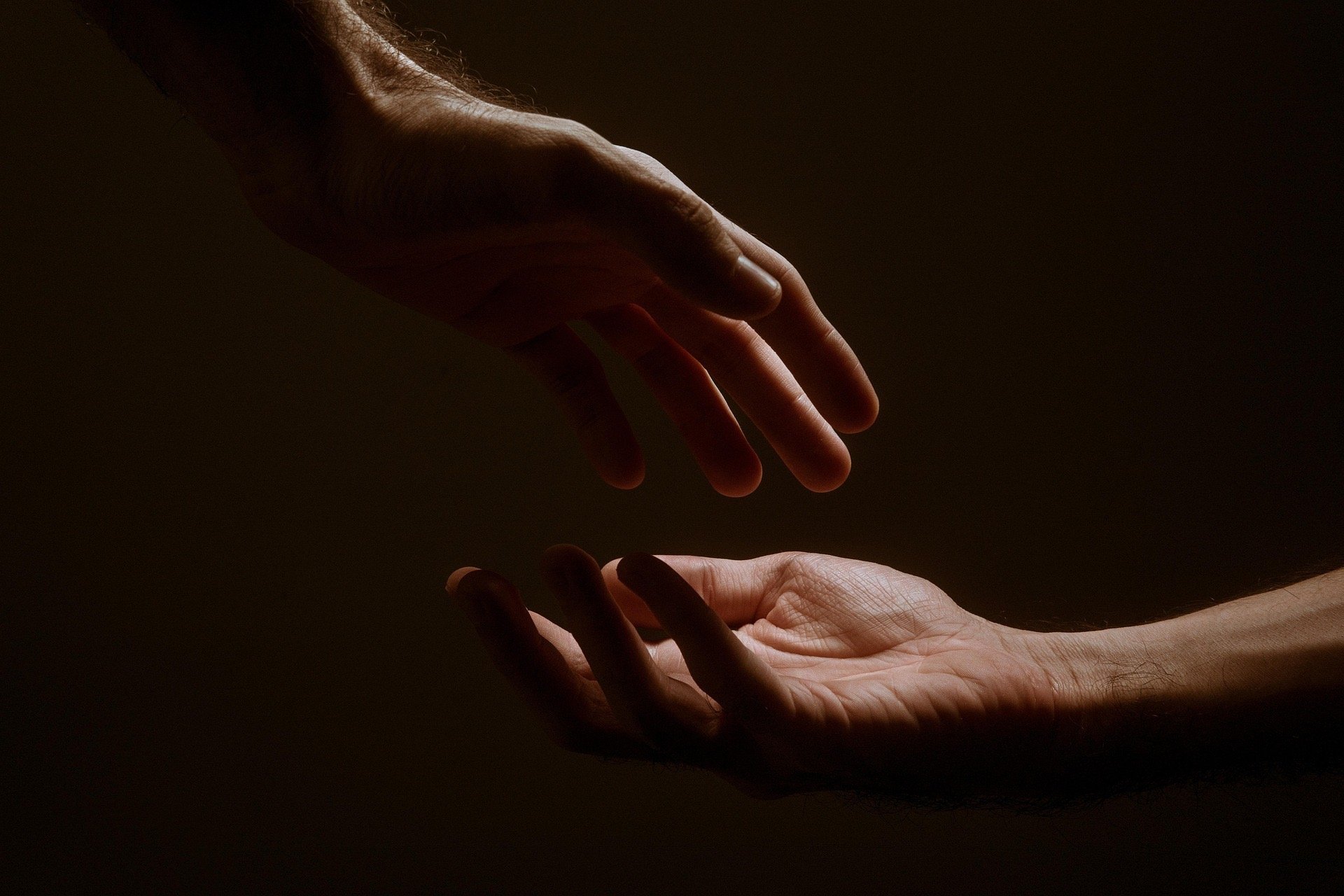 Two hands reach out towards each other against a dark background. One hand reaches from the top left corner with fingers gently curled and extended downwards. The other hand reaches from the bottom left with palm facing upwards.