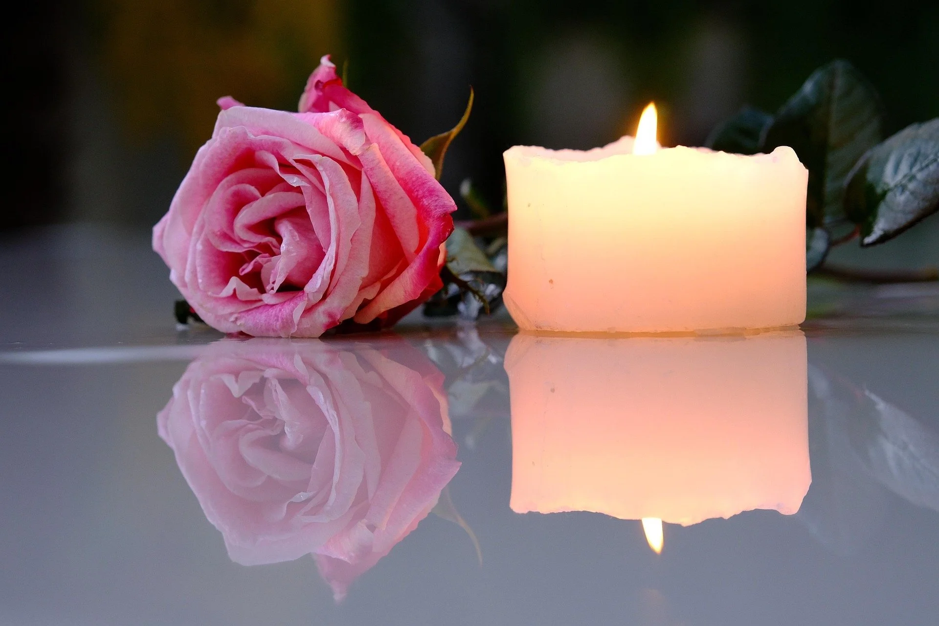A pink rose and a small lit white candle rest on a reflective surface.