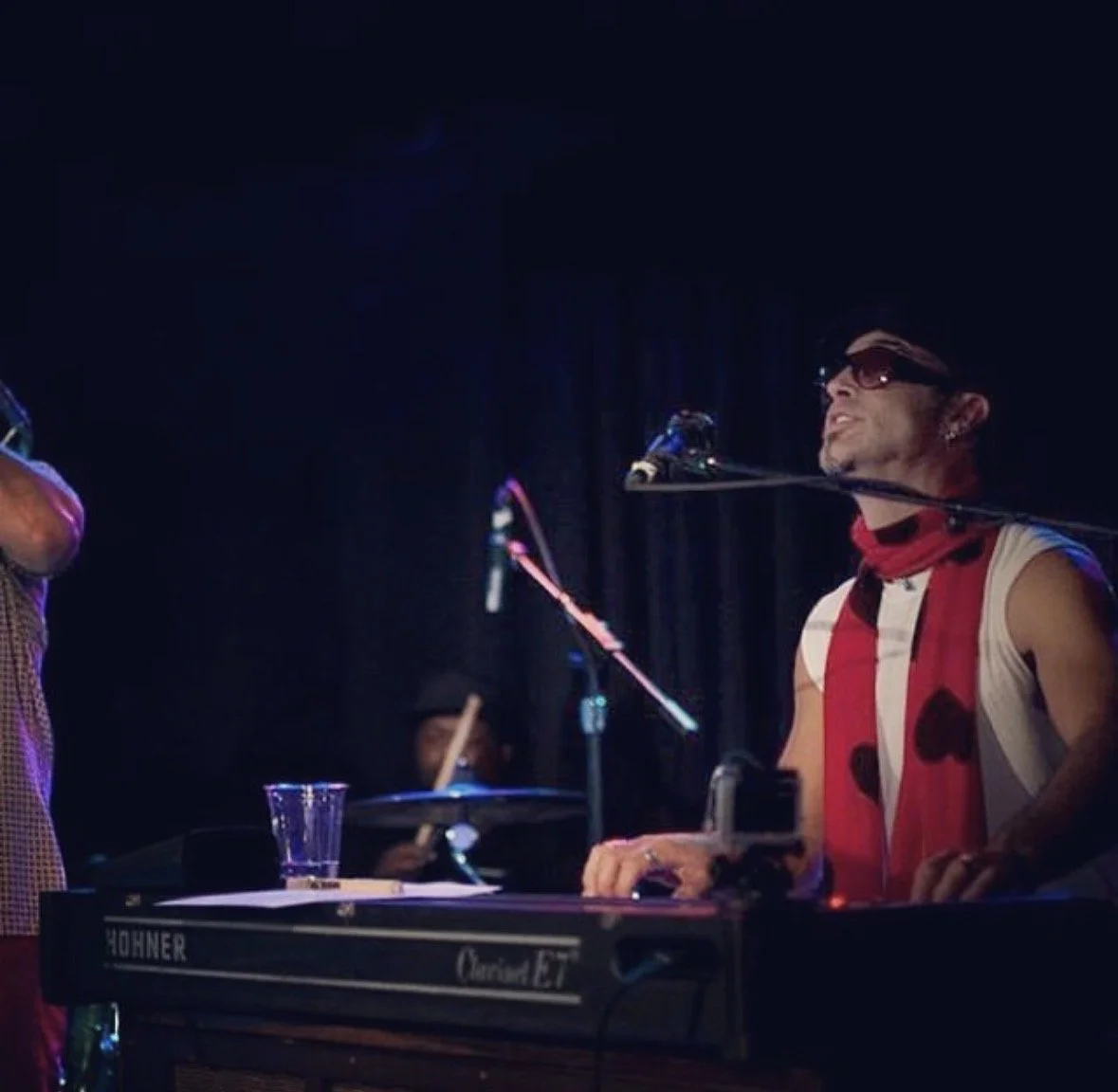 Musician wearing sunglasses and a sleeveless shirt singing into a microphone while playing a keyboard on stage.