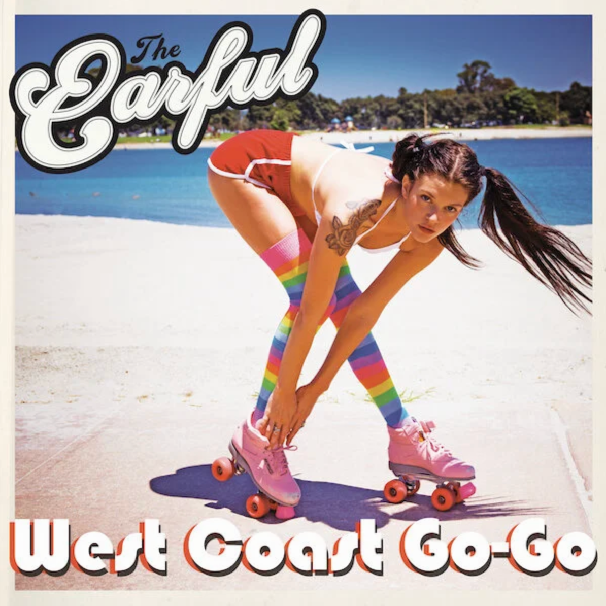 A woman roller skating on a beach with rainbow-colored knee-high socks, a tattoo on her arm, and long dark hair, with a scenic water and trees background.