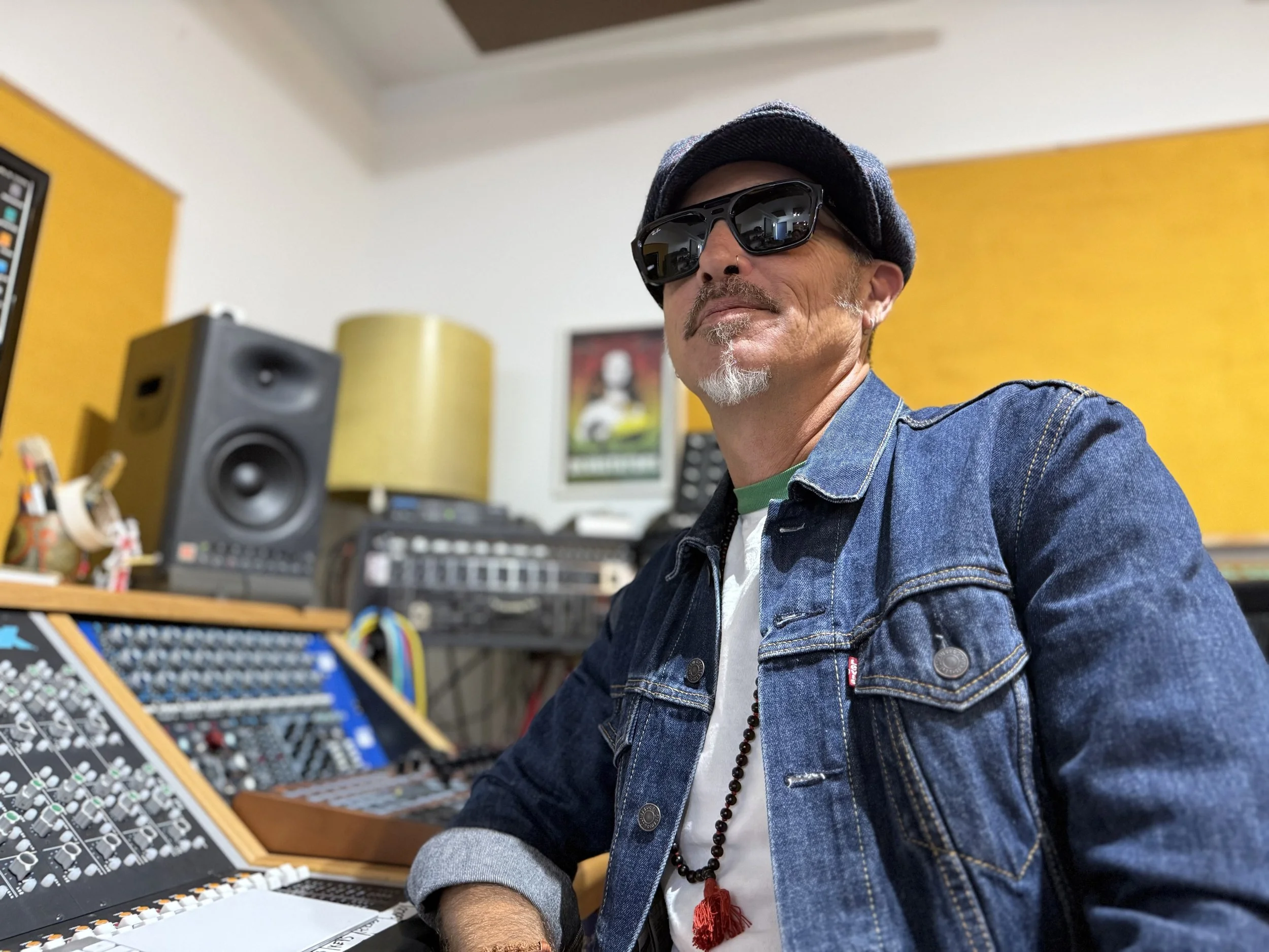 A man wearing sunglasses, a denim jacket, a black hat, and a beaded necklace sitting in a music studio with audio equipment and yellow-painted walls.