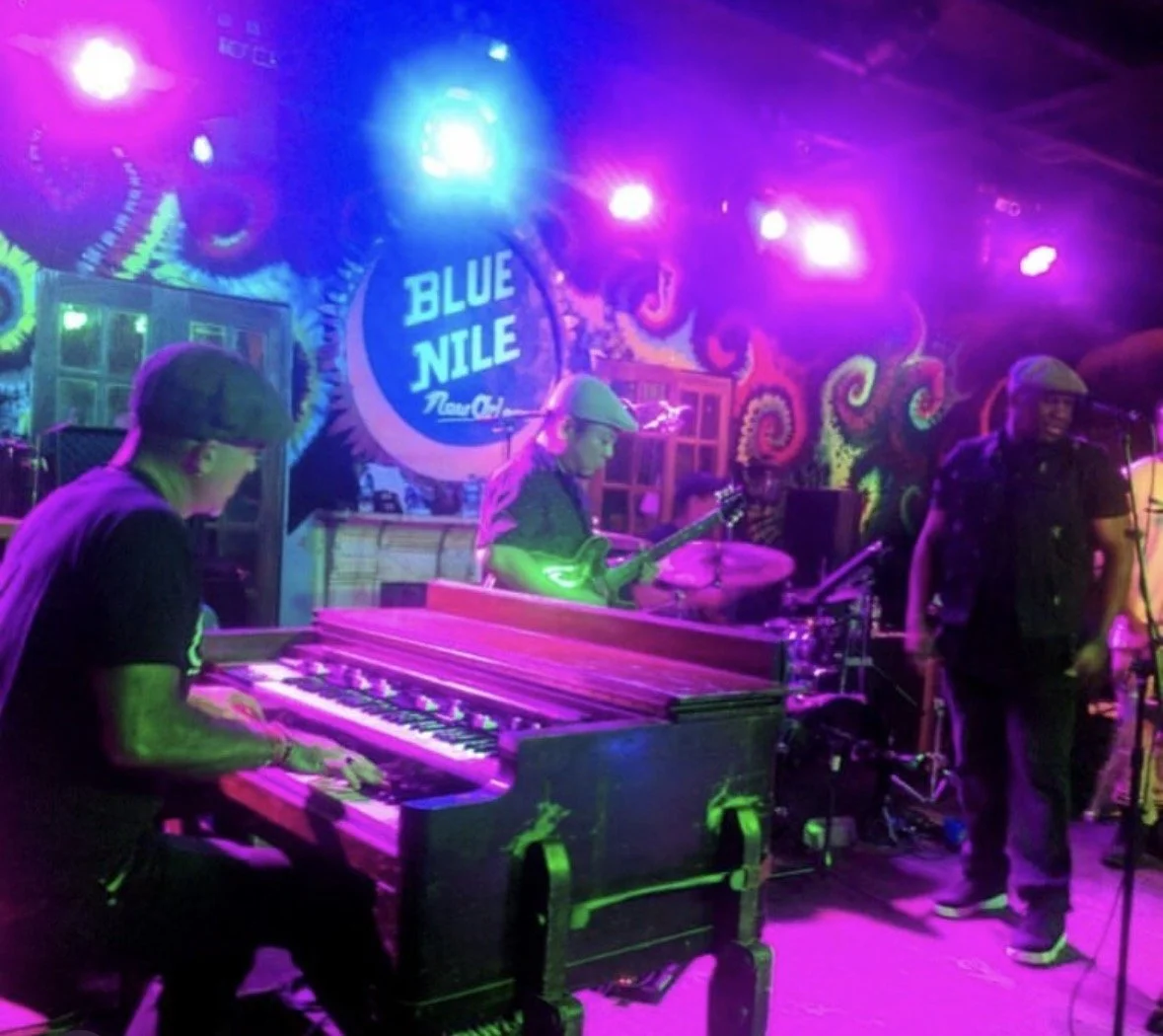 Jazz band performing on stage at Blue Nile Lounge, with pianist, guitarist, percussionist, and singer, colorful lighting, and vibrant mural backdrop.