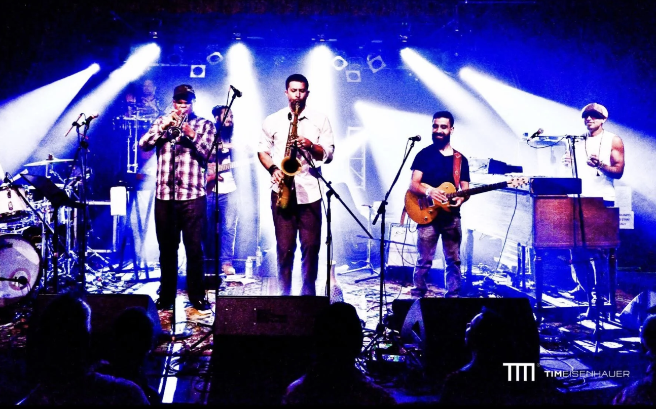 A four-piece band on stage playing instruments under blue and white spotlights, with audience members visible in the foreground. The band members include a drummer, a trumpet player, a saxophonist, a guitarist, and a keyboardist. The scene is lively with stage lighting and music equipment.