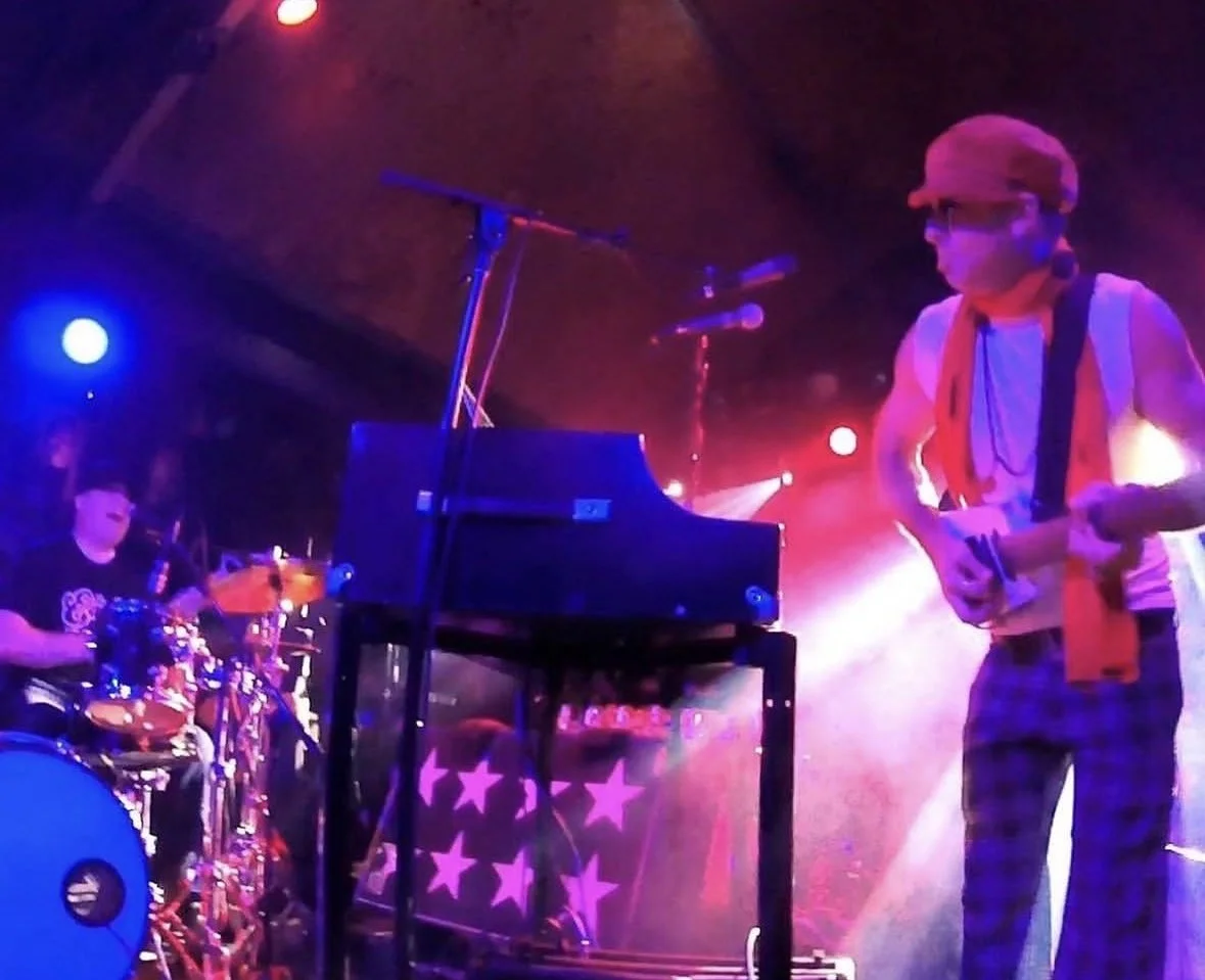 A musician playing a small guitar or ukulele on stage, wearing a red hat, sunglasses, a white sleeveless shirt, red scarf, and checkered pants, with a keyboard and a drummer in the background under colorful concert lighting.