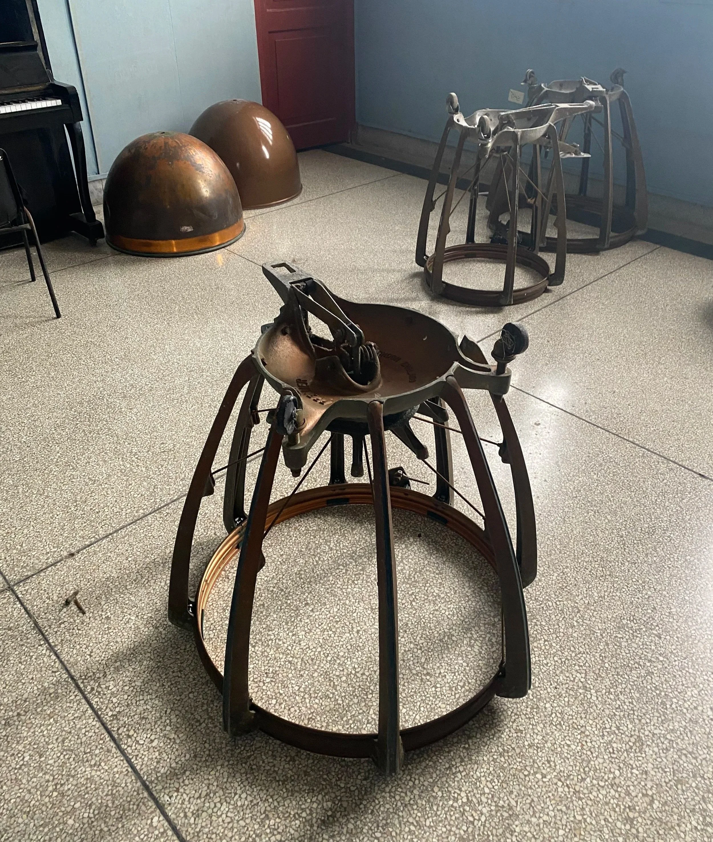 Old, rusted motorcycle frame and three rusty metal helmets on a speckled tile floor, with a blue wall and a red cabinet in the background.