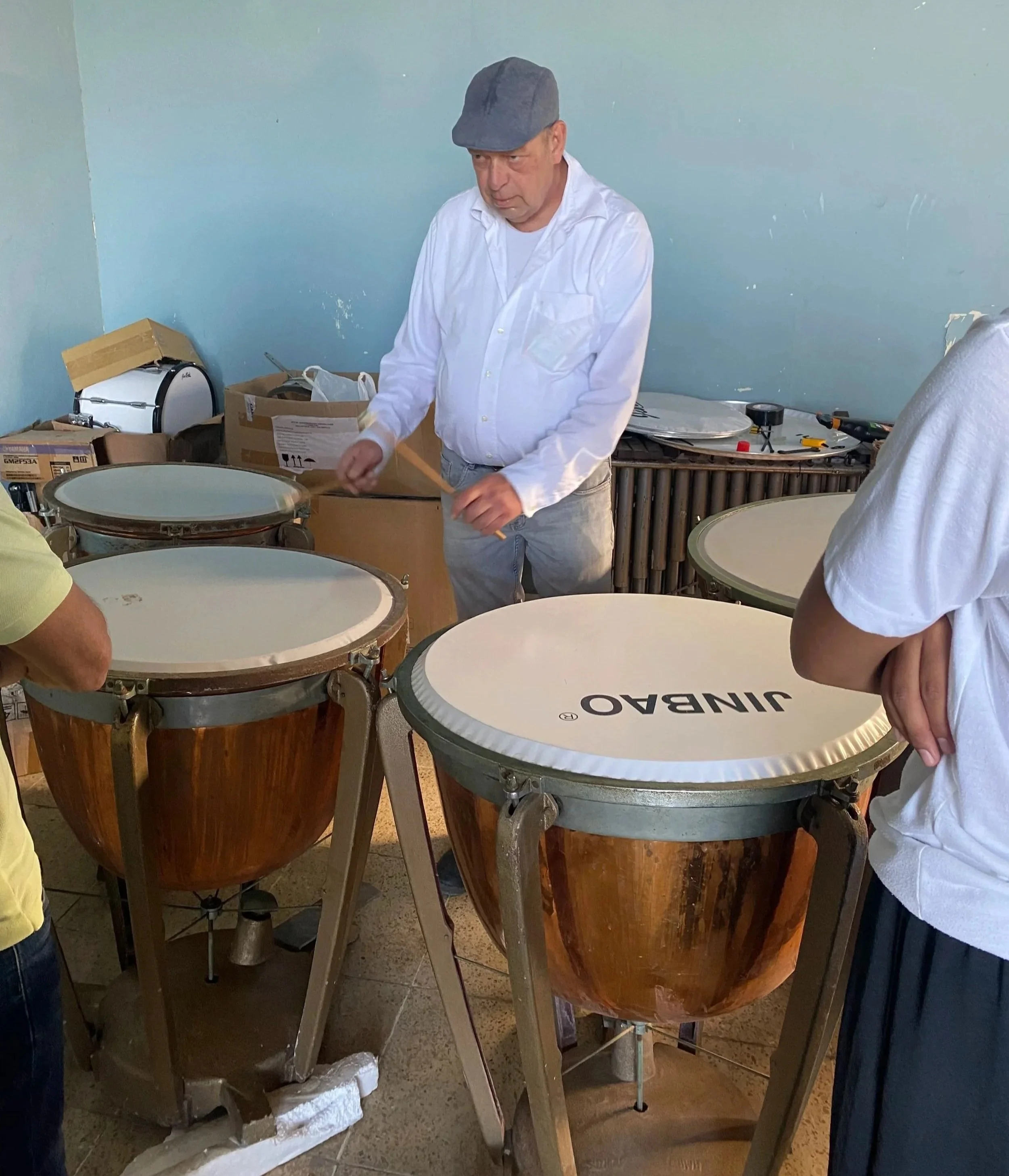 A man in a white shirt and a gray cap standing among percussion drums, including congas with a wooden finish and a drum with a white top labeled 'JINBO'. There are boxes and tools on the side, suggesting setup or maintenance of the drums, in a room w