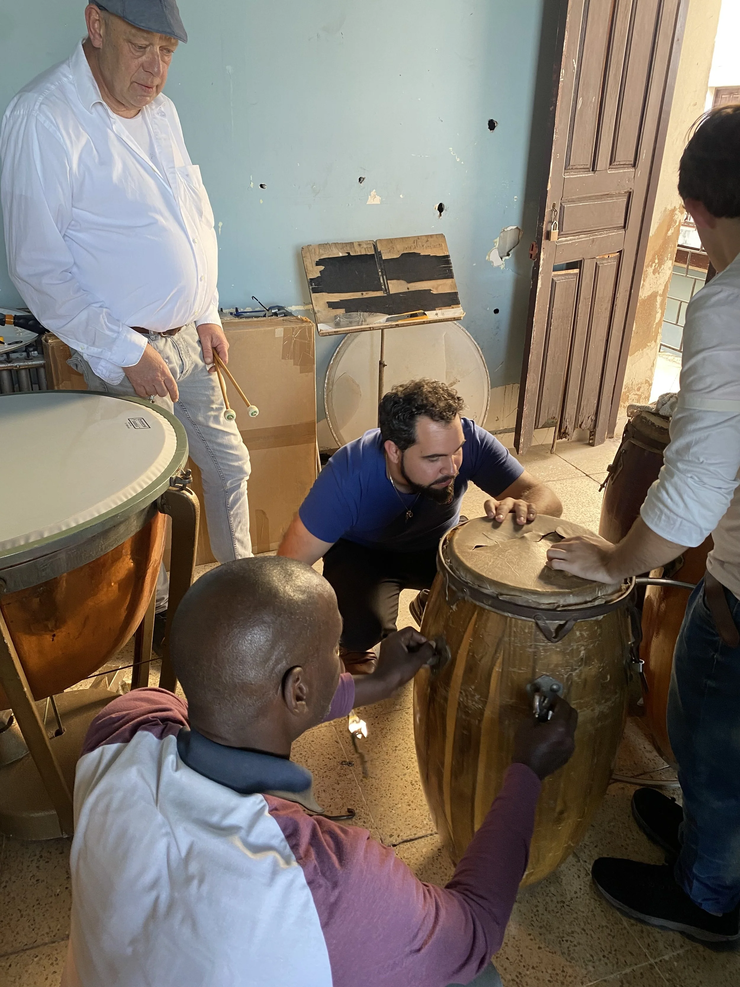 Group of people working together on traditional drums in a room with a wooden door and a damaged wall.