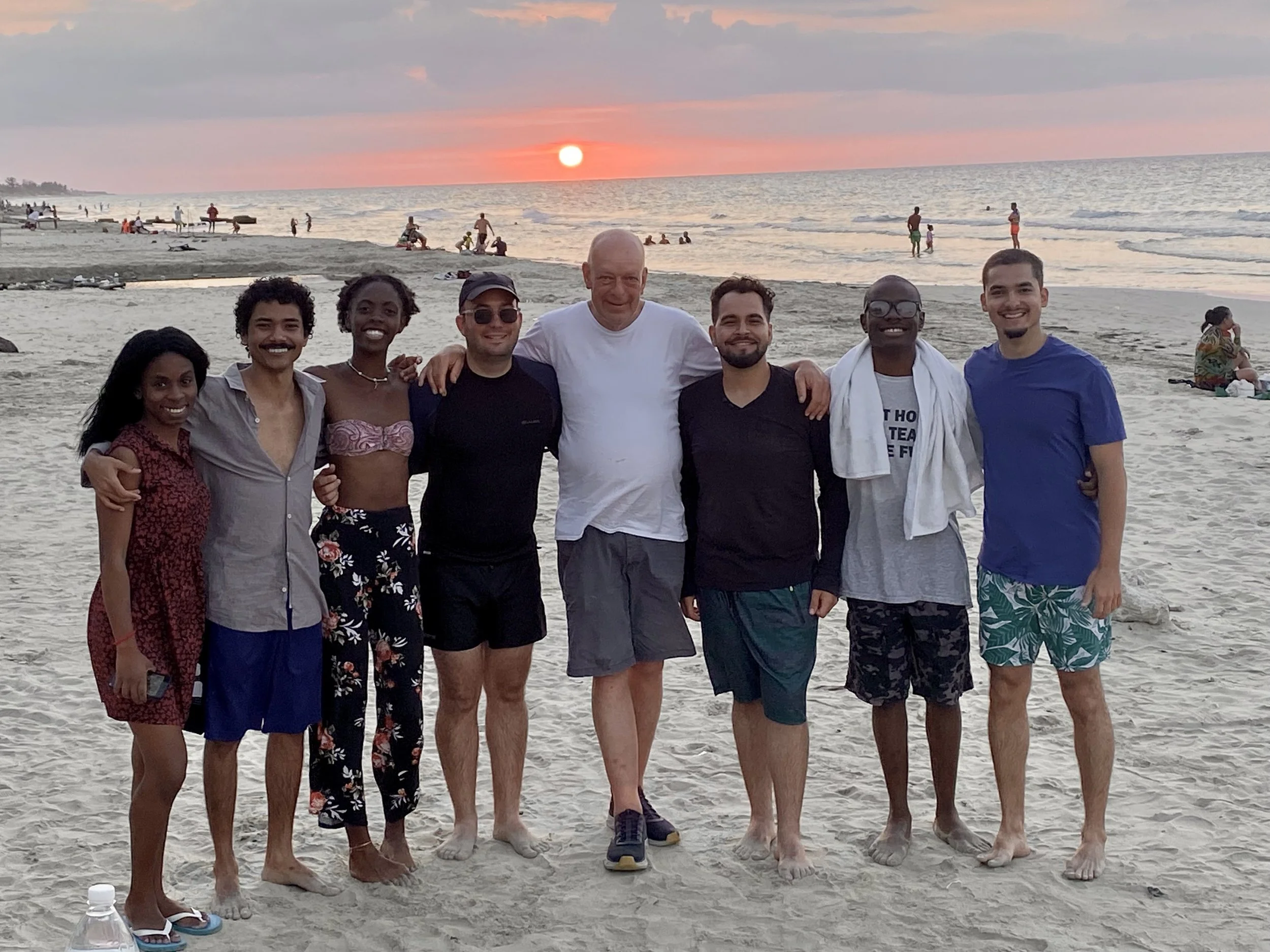 Group of eight people standing on the beach at sunset, smiling and posing for a photo.