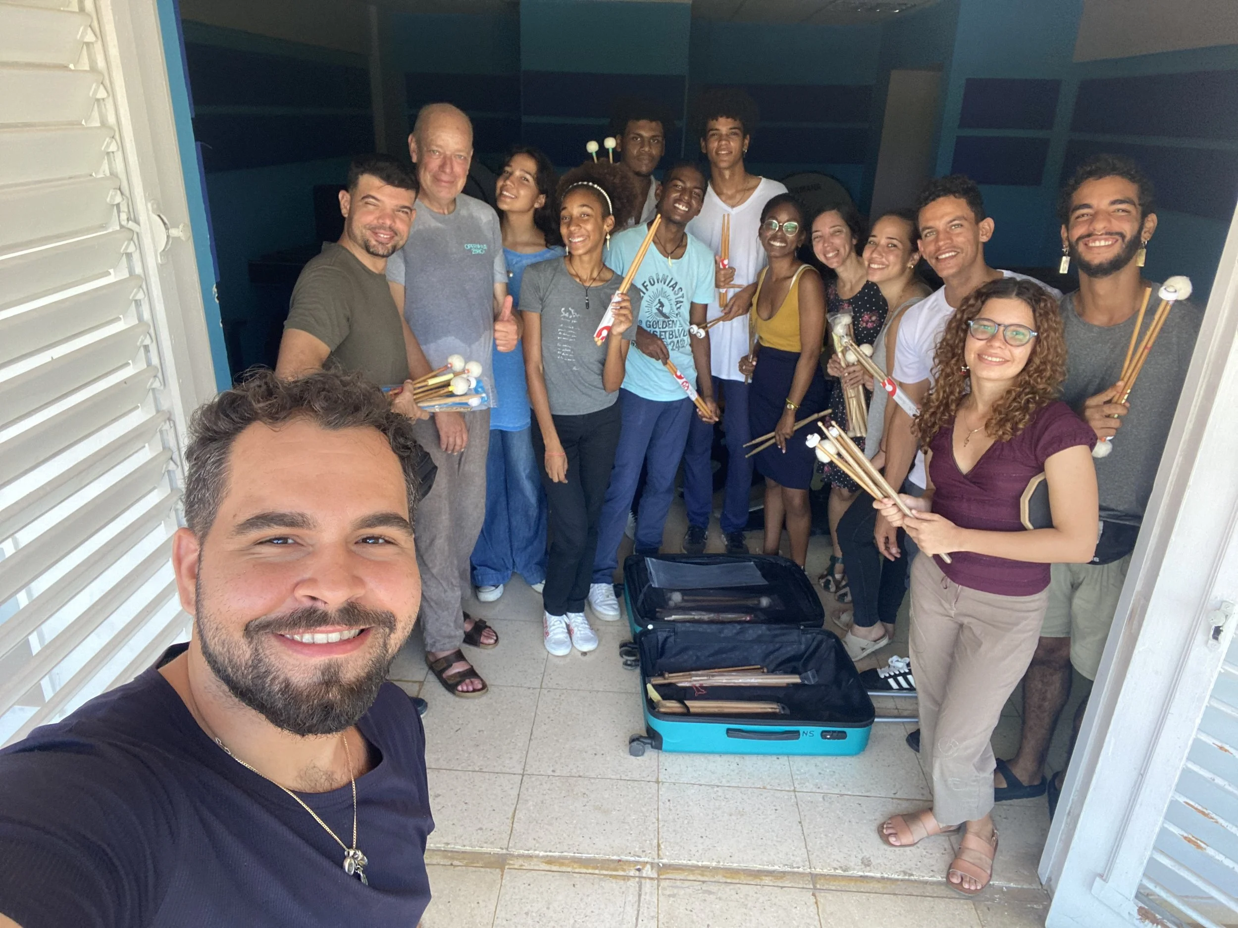 Group of people standing together indoors, holding drumsticks, smiling for a photo, with an open suitcase on the floor.