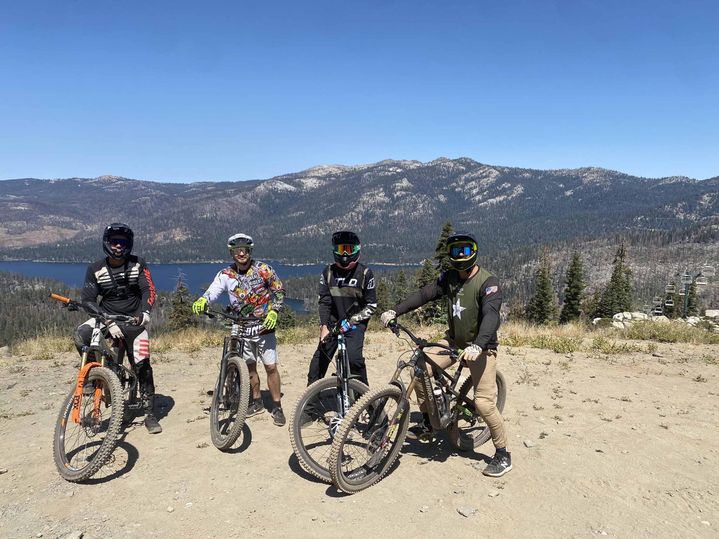 Four mountain bikers wearing helmets and goggles standing with their bikes on a dirt trail with a mountain lake and forested mountains in the background under a clear blue sky.