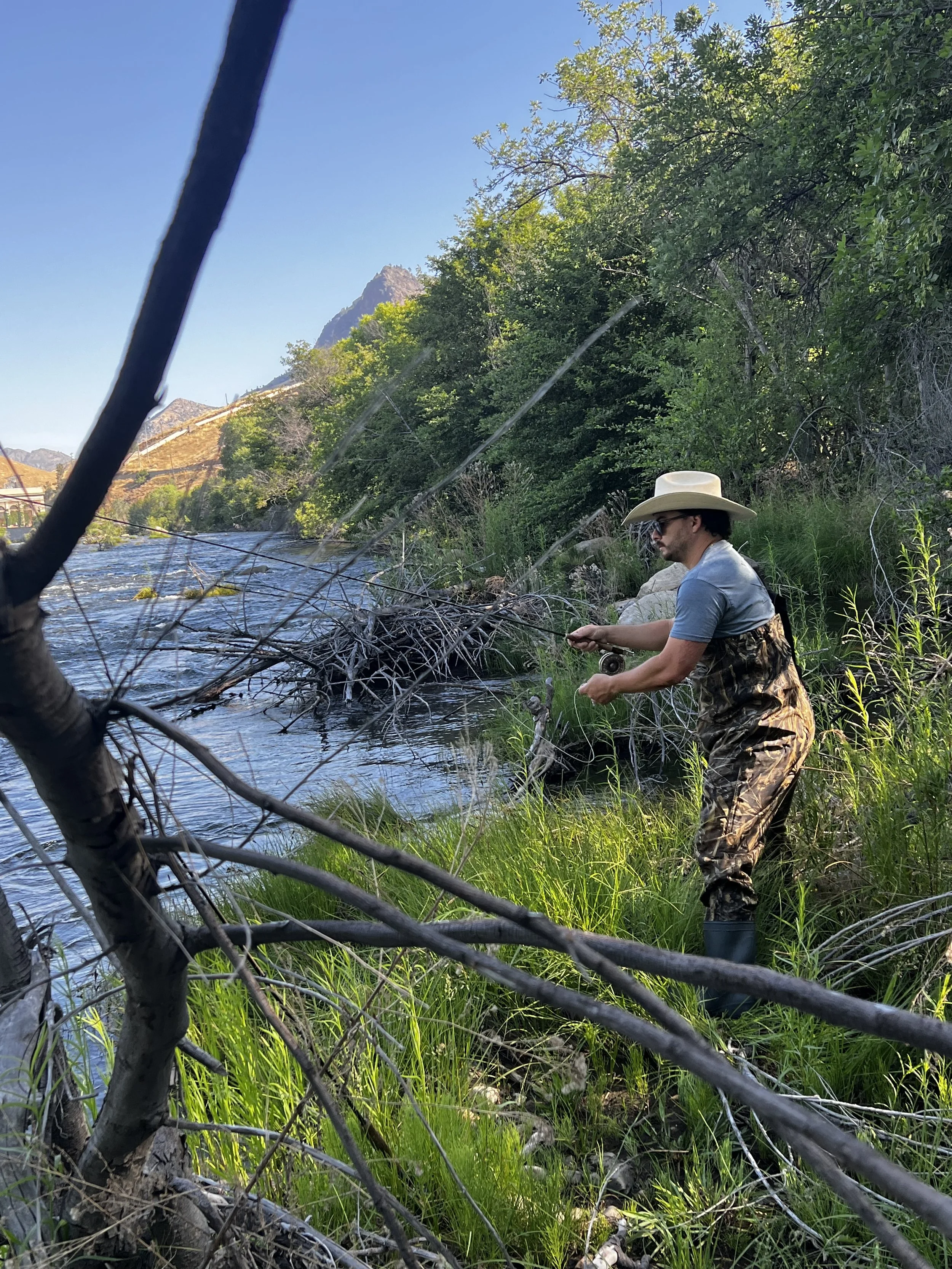 A man wearing a large white hat, sunglasses, a gray shirt, and camouflage overalls fishing beside a river with greenery and mountains in the background.