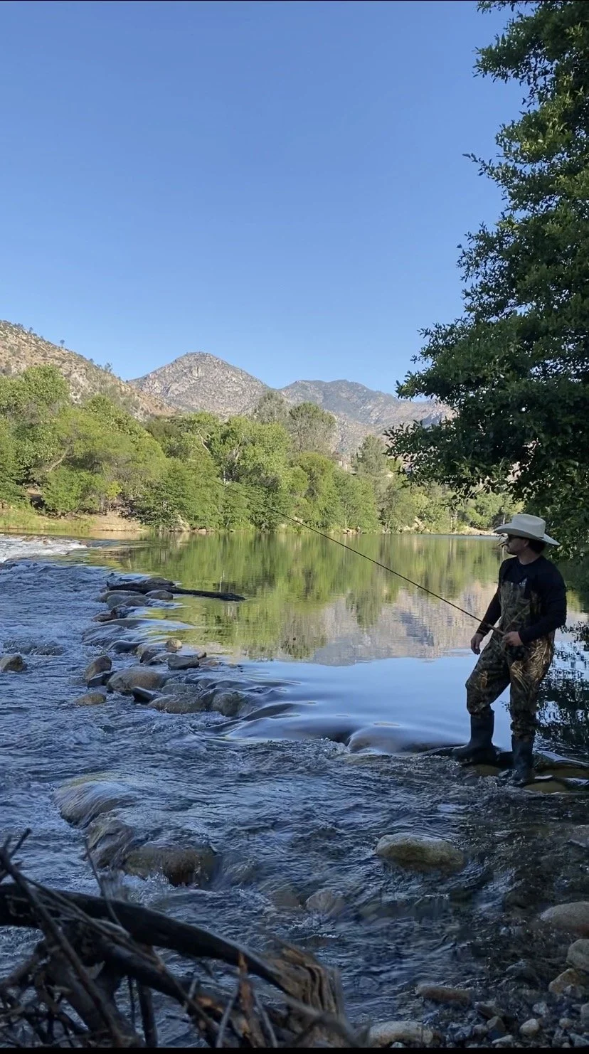 A man fishing in a river with mountains and green trees in the background on a clear day.