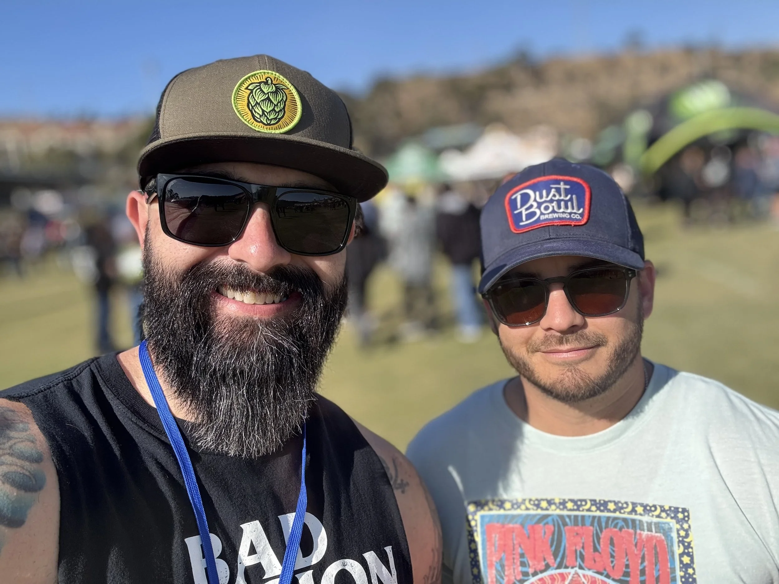 Close-up of two smiling men wearing sunglasses and caps at an outdoor event. The man on the left has a beard, wears a black shirt, and a cap with a green hop cone logo. The man on the right is clean-shaven, wears a light-colored shirt, and a cap with a