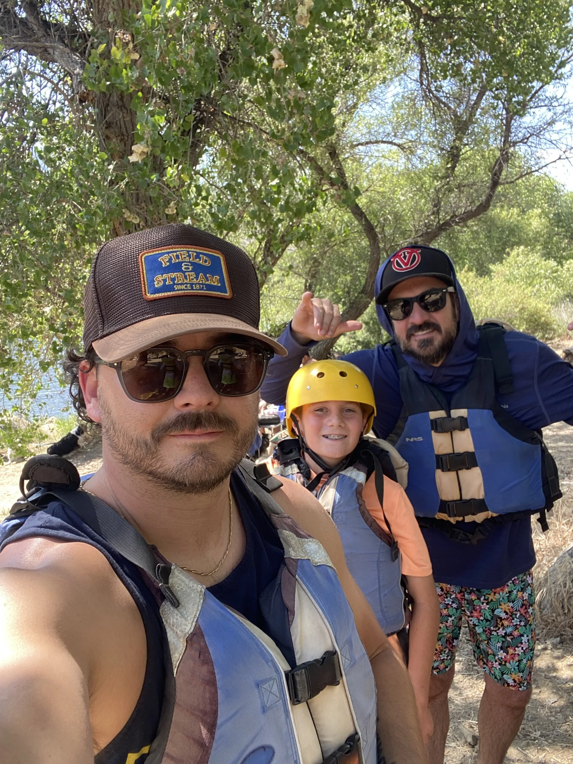 Four people enjoying an outdoor activity under trees, wearing helmets and casual summer clothing, with a background of trees and a clear sky.