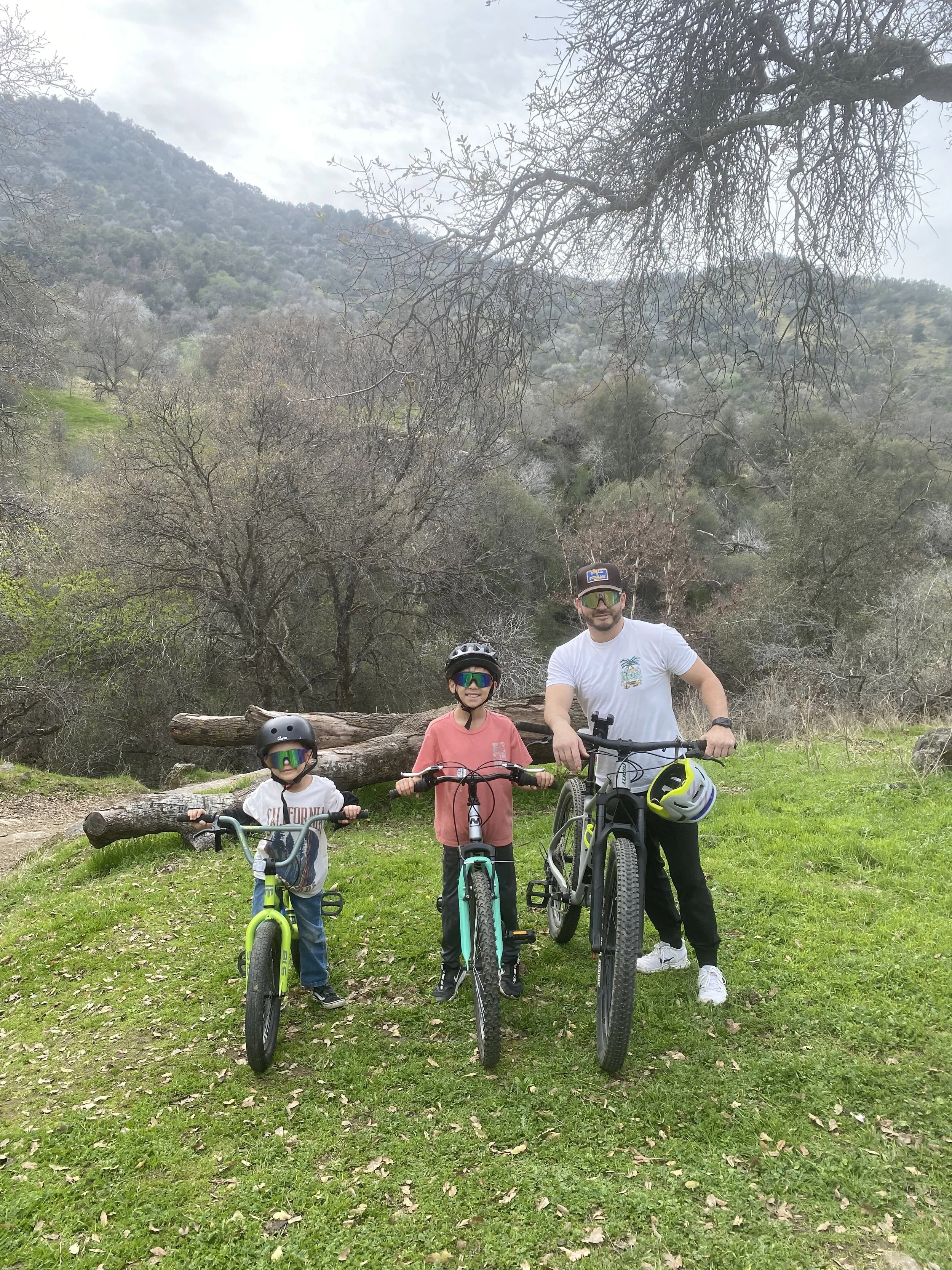 A family of three hiking with bikes in a green outdoor area with trees and hills in the background, under a cloudy sky.