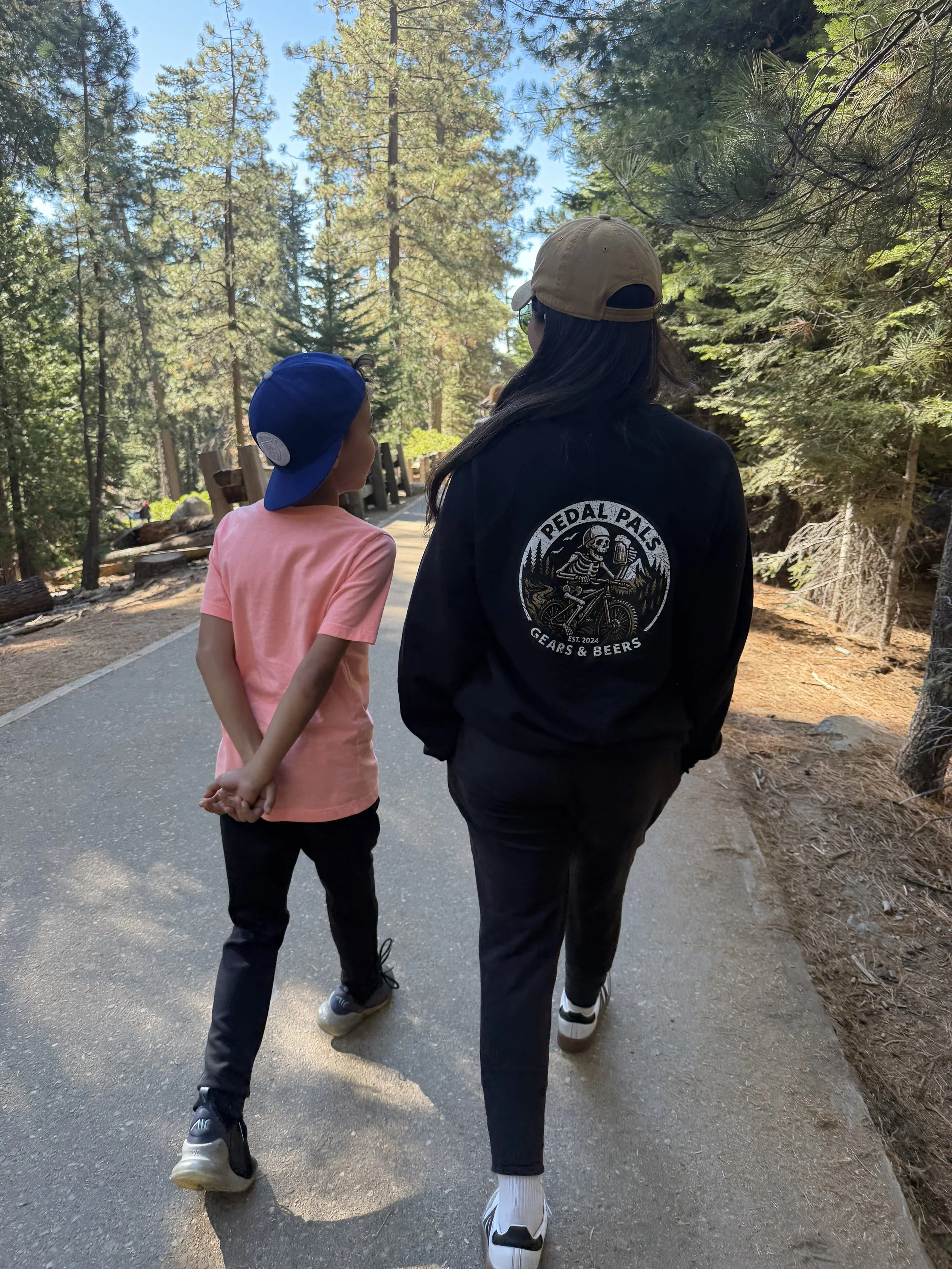 A woman and a young girl walk together on a trail in a forested area during daytime, holding hands behind their backs.