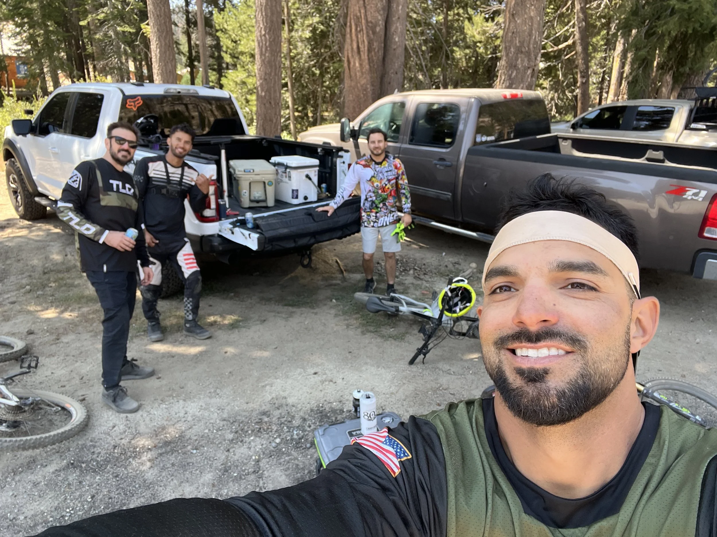 Group of four men with mountain bikes and gear in a forested area, some smiling and posing for a photo, with trucks and trees in the background.