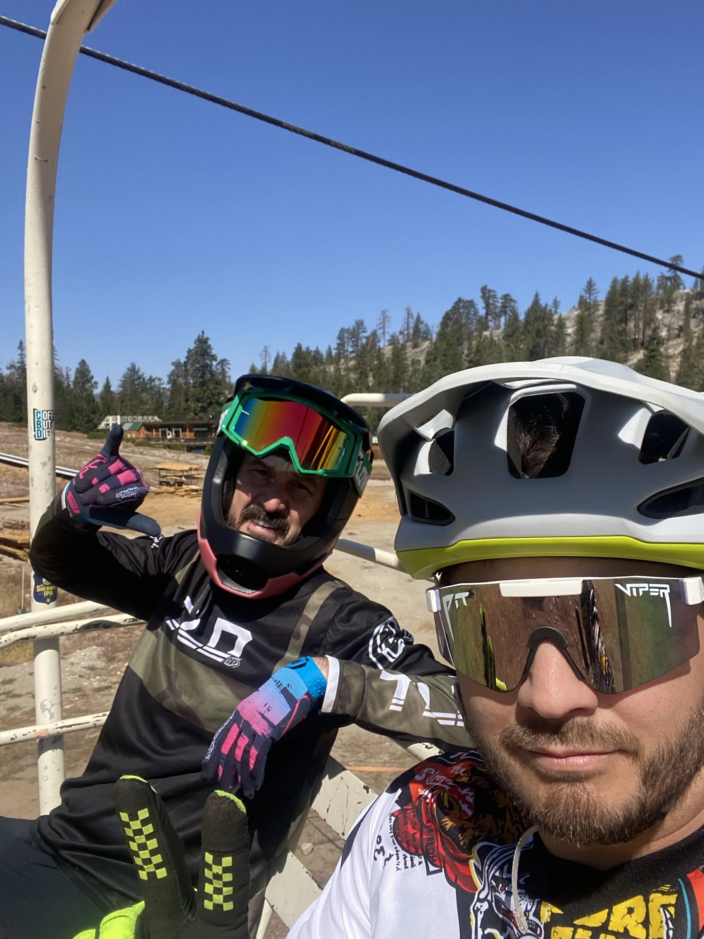 Two male mountain bikers wearing helmets and goggles, taking a selfie outdoors near a ski lift with a cable in the background, trees, and blue sky.