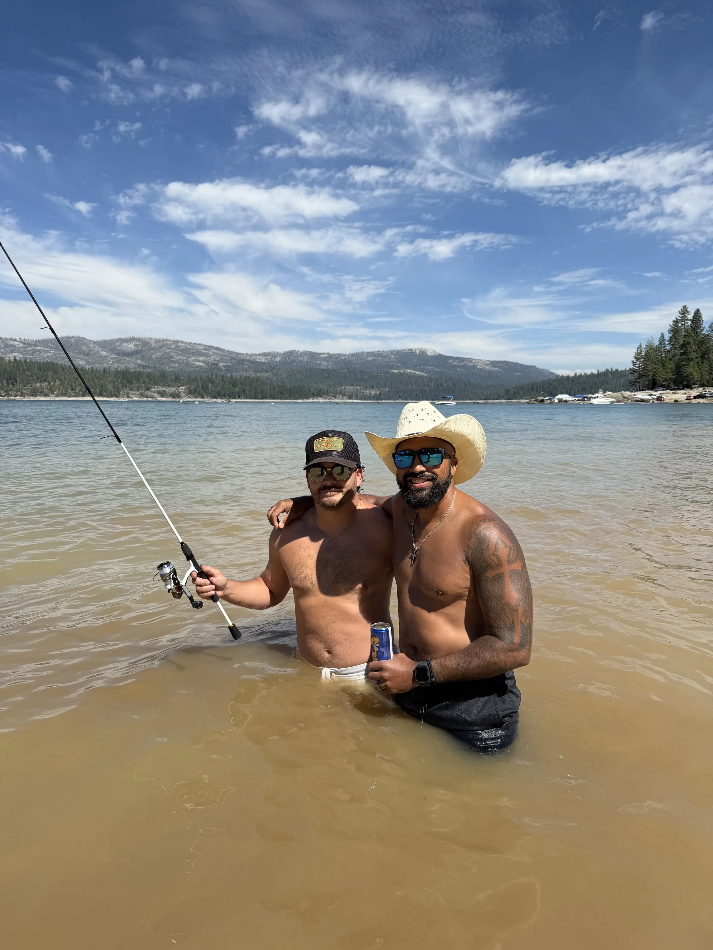 Two men are standing in shallow water at the beach, smiling at the camera. One man is wearing a black hat, sunglasses, and holding a fishing rod. The other man is wearing a cowboy hat, sunglasses, and holding a can. The background features mountains, a forested shoreline, and a blue sky with scattered clouds.
