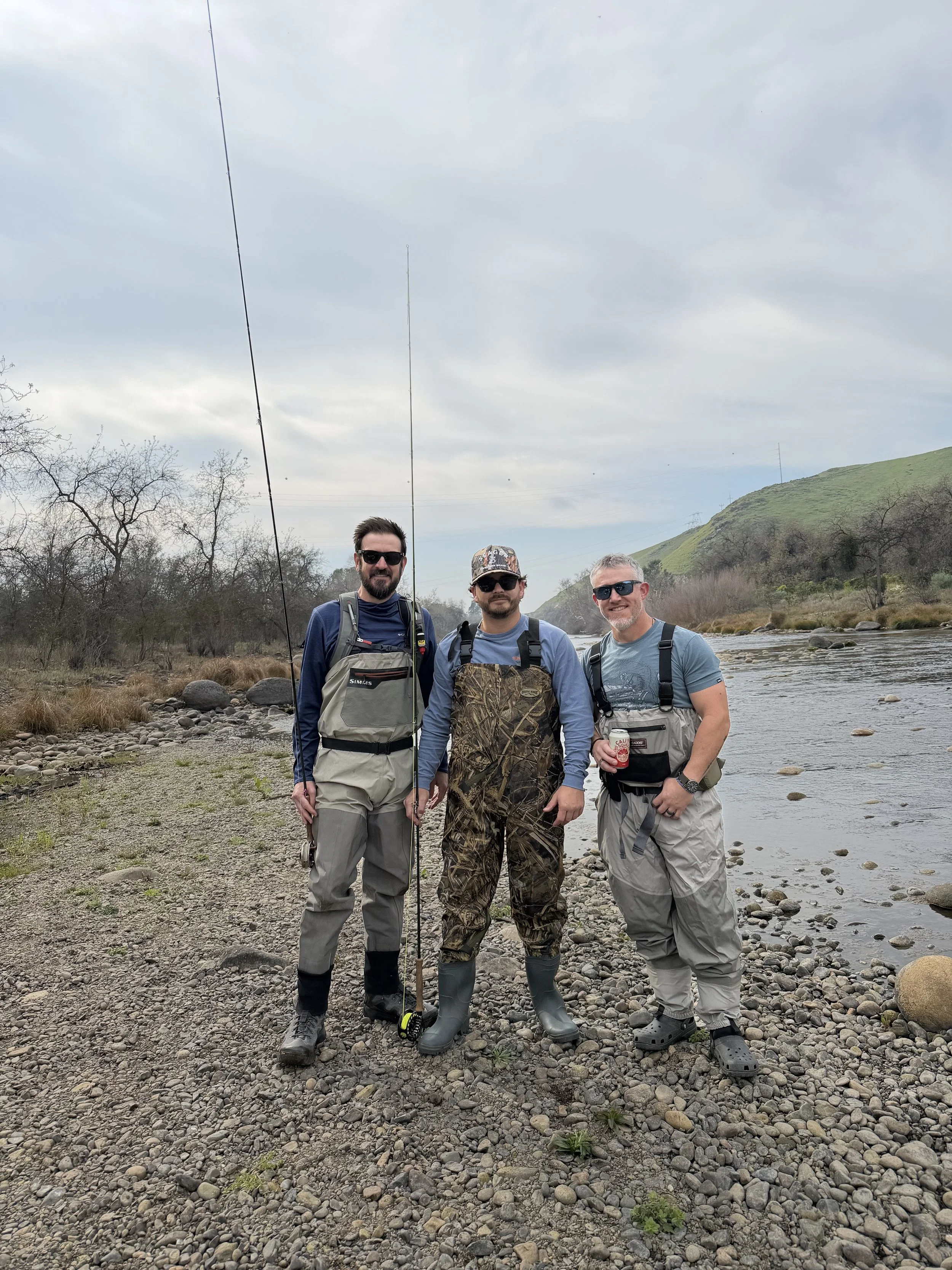 Three men standing on a rocky riverbank holding fishing gear, wearing outdoor clothing, with hills, trees, and a cloudy sky in the background.